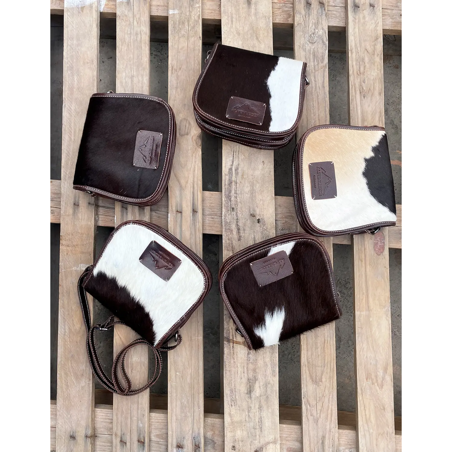 Fünf exquisite Umhängetaschen VRONI mit Rindsledermustern in Schwarz, Braun und Weiß sind kunstvoll auf einer Holzpalette präsentiert. Jede Tasche besticht durch ein einzigartiges Farbschema und einen kleinen Patch mit dem Logo.