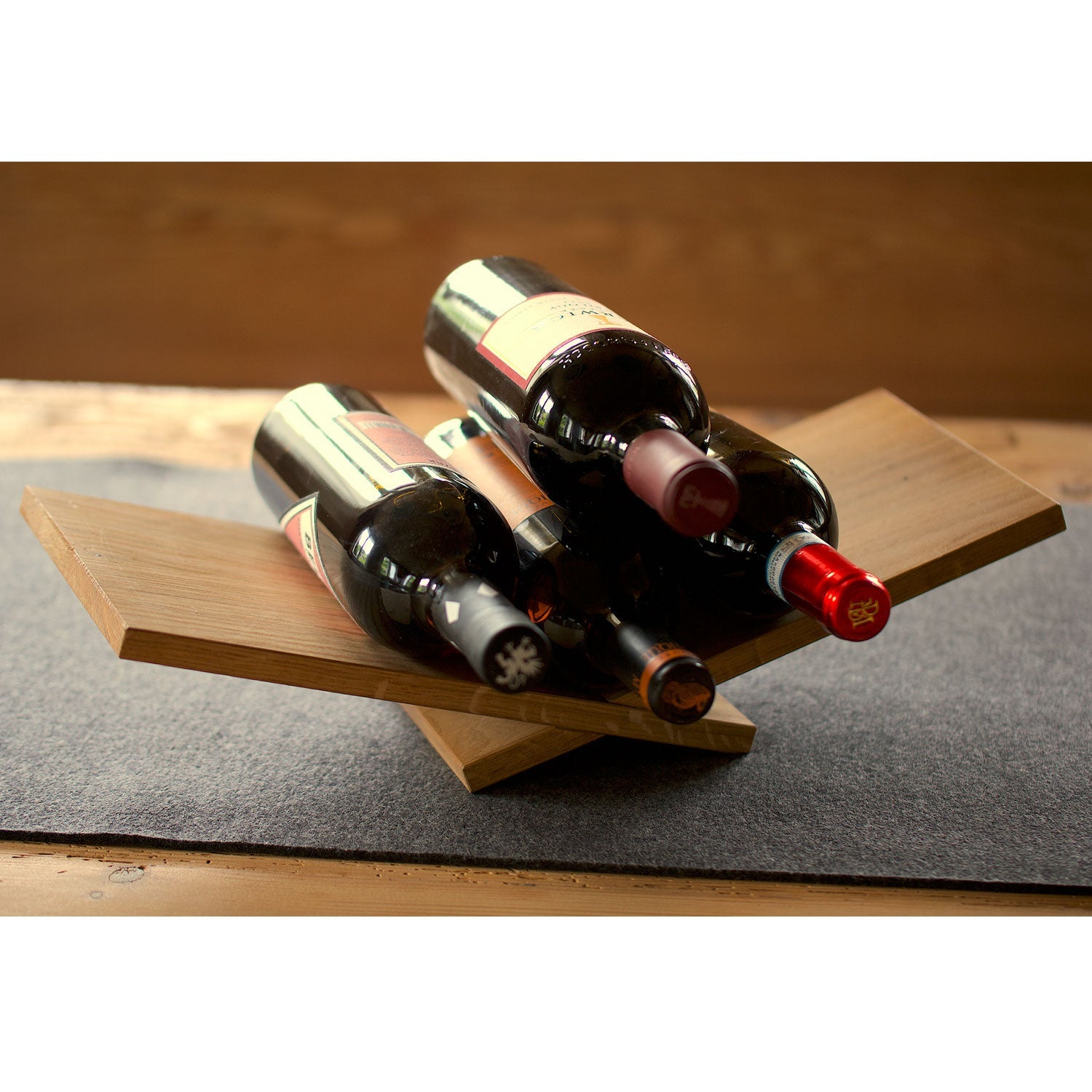 Die Weinliege ist ein modernes, X-förmiges Weinregal aus Holz, in dem fünf Flaschen horizontal auf einer Holzoberfläche gelagert werden können. Das minimalistische Design wird durch ein graues Tischset ergänzt und verbindet auf elegante Weise Form und Funktion.