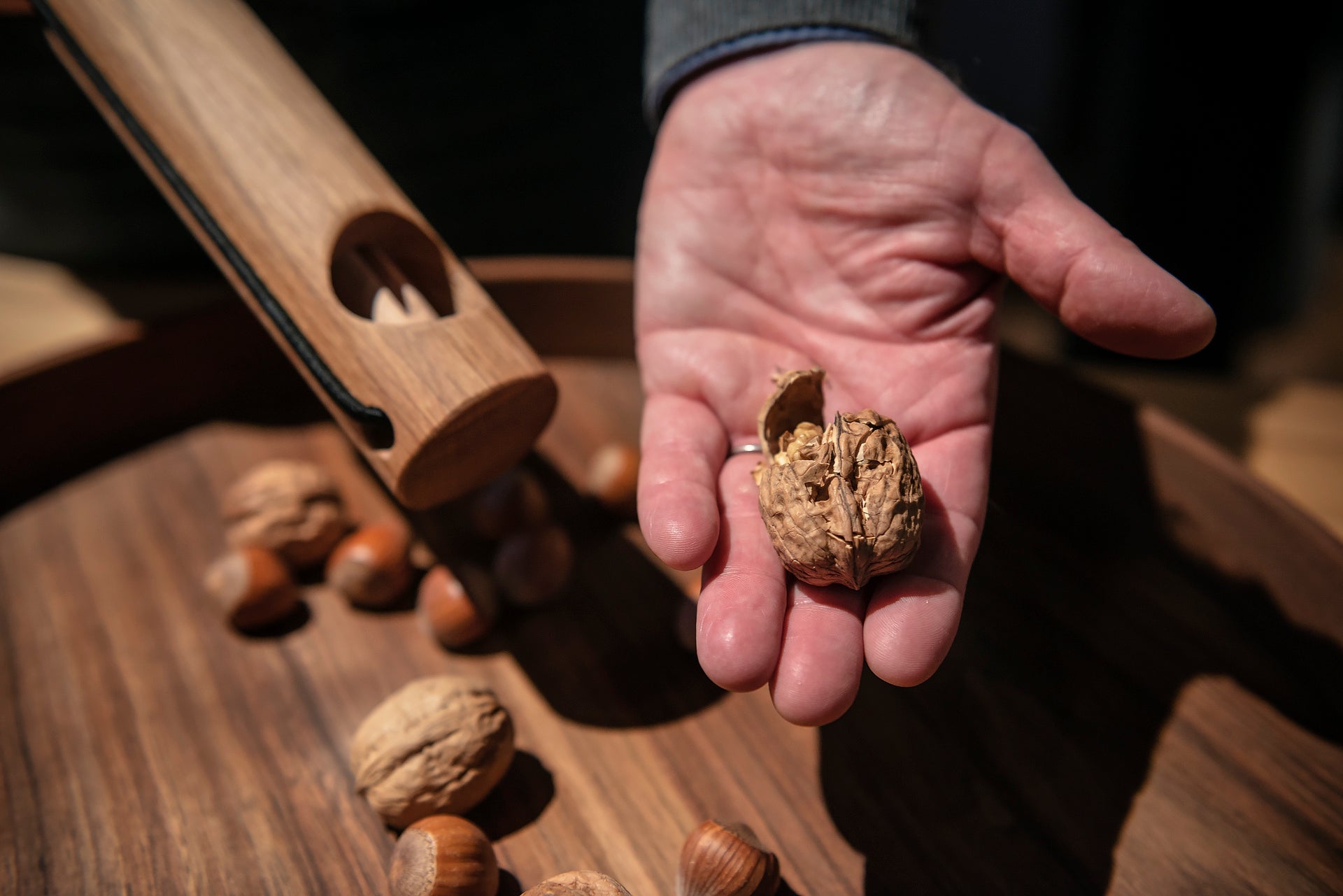 Eine Hand hält eine geknackte Walnuss neben einem Nussknacker und gemischten Nüssen auf einem Eichentablett, inspiriert von Dietmar Mechsner.