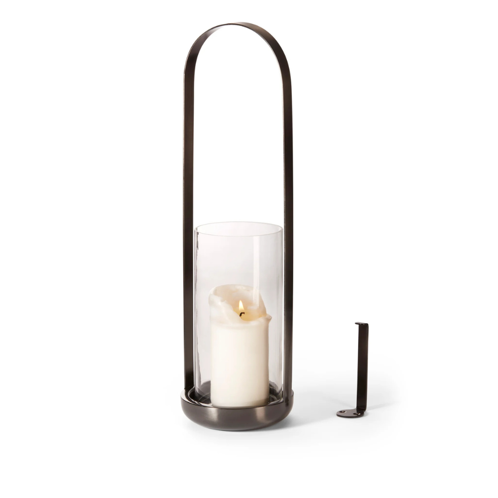 Im Windlicht CALA, einer Laterne aus Glas und Metall mit einem Glaszylinder, brennt eine weiße Kerze, daneben befindet sich eine passende Wandhalterung – perfekt für einen Hauch romantischer Reminiszenz.