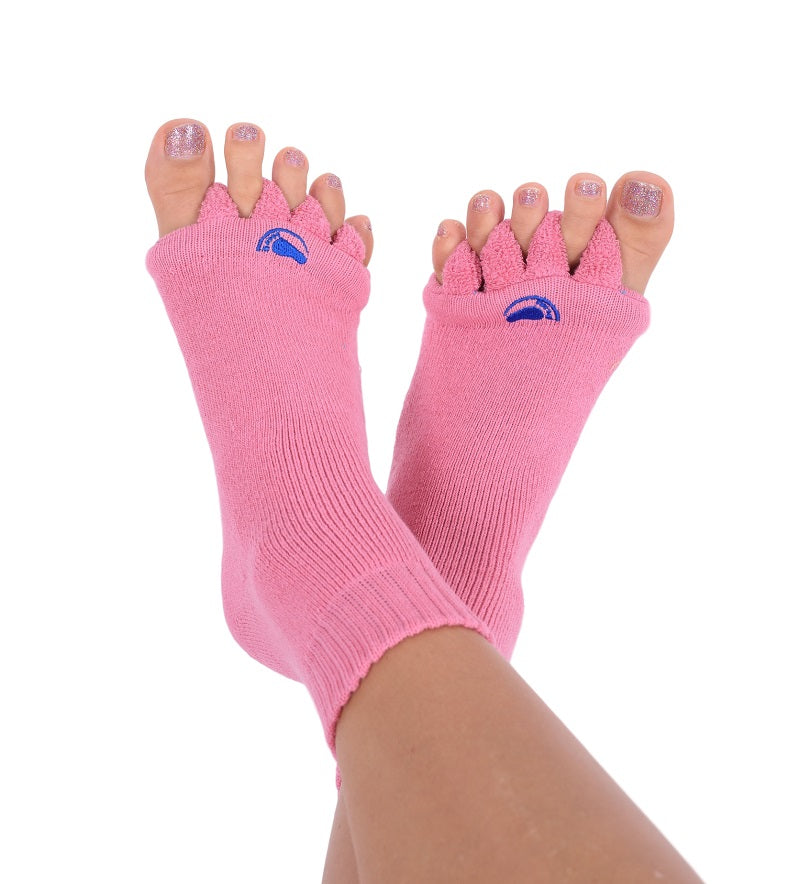 Zwei Füße tragen pinkfarbene, offene Fußausrichtungssocken für die Pediküre. Jeder Zeh ist separat und gibt den Blick auf glitzernden Nagellack frei. Diese bequemen Socken präsentieren sich mit cleverer Zehenspreizer-Funktionalität auf schlichtem Hintergrund und betonen so Stil und Fußausrichtung.