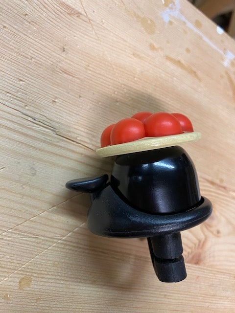 Die Schwarzwald Bollenhut Fahrradklingel, eine schwarze Fahrradklingel aus Kunststoff mit roten und beigen Details, sitzt auf einer hellen Holzoberfläche und bringt einen Hauch Schwarzwald-Flair auf Ihre Fahrt.