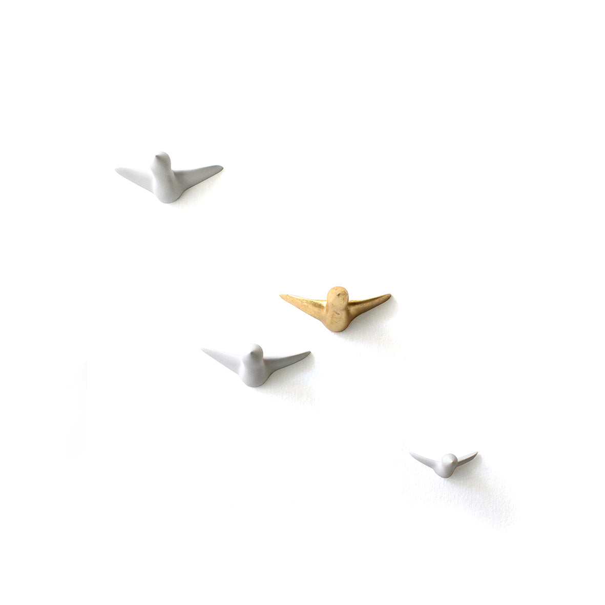 Die Wandskulptur - kleiner Vogelschwarm, bestehend aus vier minimalistischen Vogelskulpturen (drei weiße, eine goldene), wird mit Wandhaken an einer schlichten weißen Wand befestigt. Die Vögel sind in unterschiedlichen Flugrichtungen angeordnet, wodurch ein funktionales und elegantes Design entsteht.