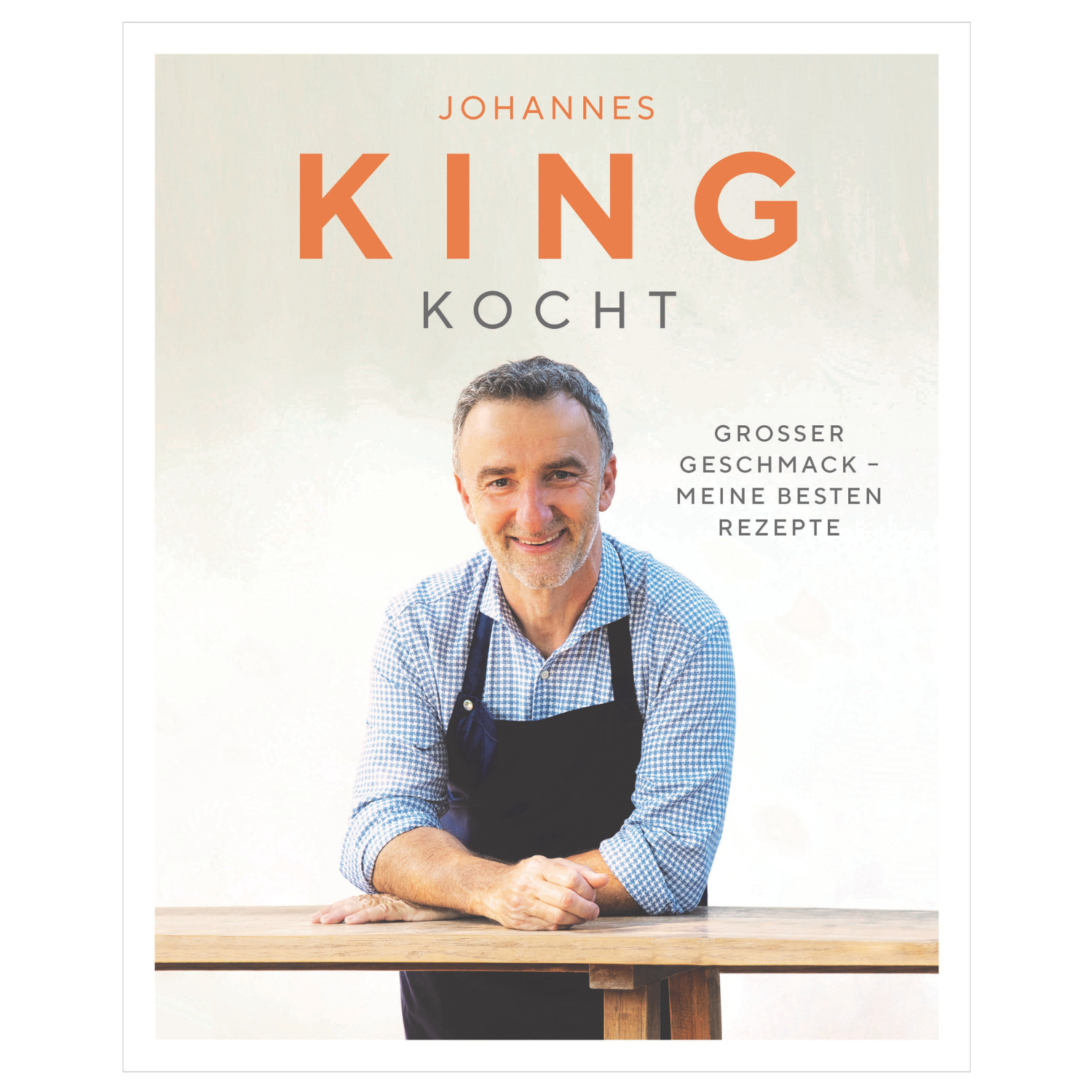 Ein lächelnder Mann in einer Schürze lehnt an einem Tisch; Text lautet King kocht – Sternekoch Johannes King präsentiert Genussfreude und kreative Rezepte im deutschen Rezeptbuch.