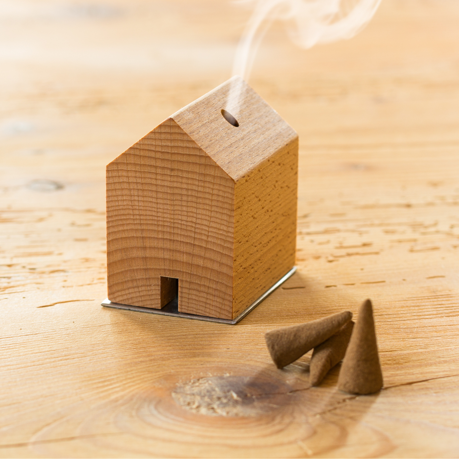 Das Räuchermännchen Rauchhaus ist ein kleines Räuchergefäß aus Holz, aus dem Rauch aufsteigt, der neben drei braunen Räucherkegeln auf einer Holzoberfläche präsentiert wird.