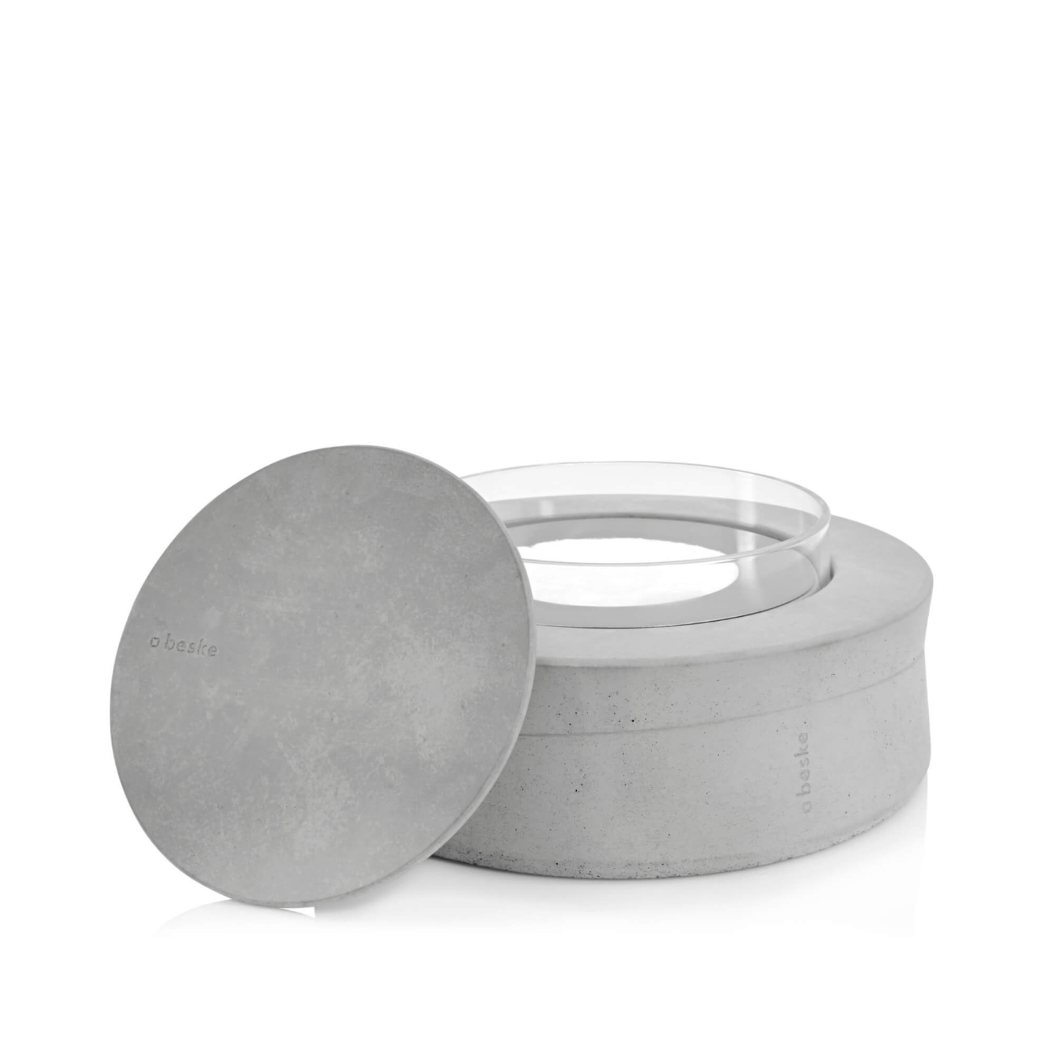 Das Ethanolfeuer – Beske Manufaktur ist ein runder Betonbehälter mit einem Glaskern und passendem flachen Deckel, beide mit der Aufschrift „Basie“ versehen – ideal als stilvoller Tischkamin für B-Ethanolfeuer.