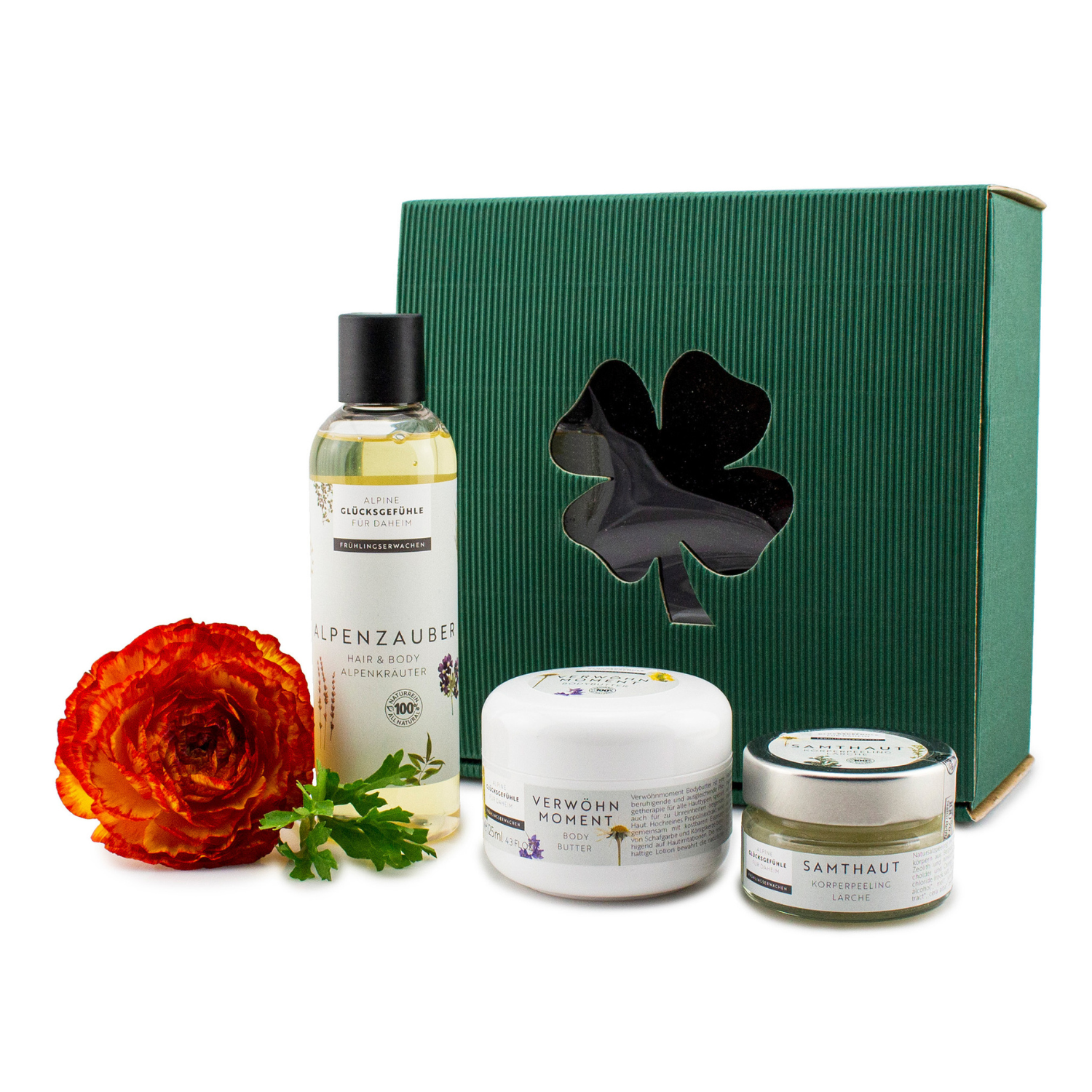Geschenkset „Zauberkraft“: Grünes Geschenkbox mit Alpenzauber Pflege, rot-orangefarbener Blume und Kleeblatt-Ausschnitt. Enthält Alpenkräuter Körperpeeling für ein belebendes Verwöhnerlebnis.