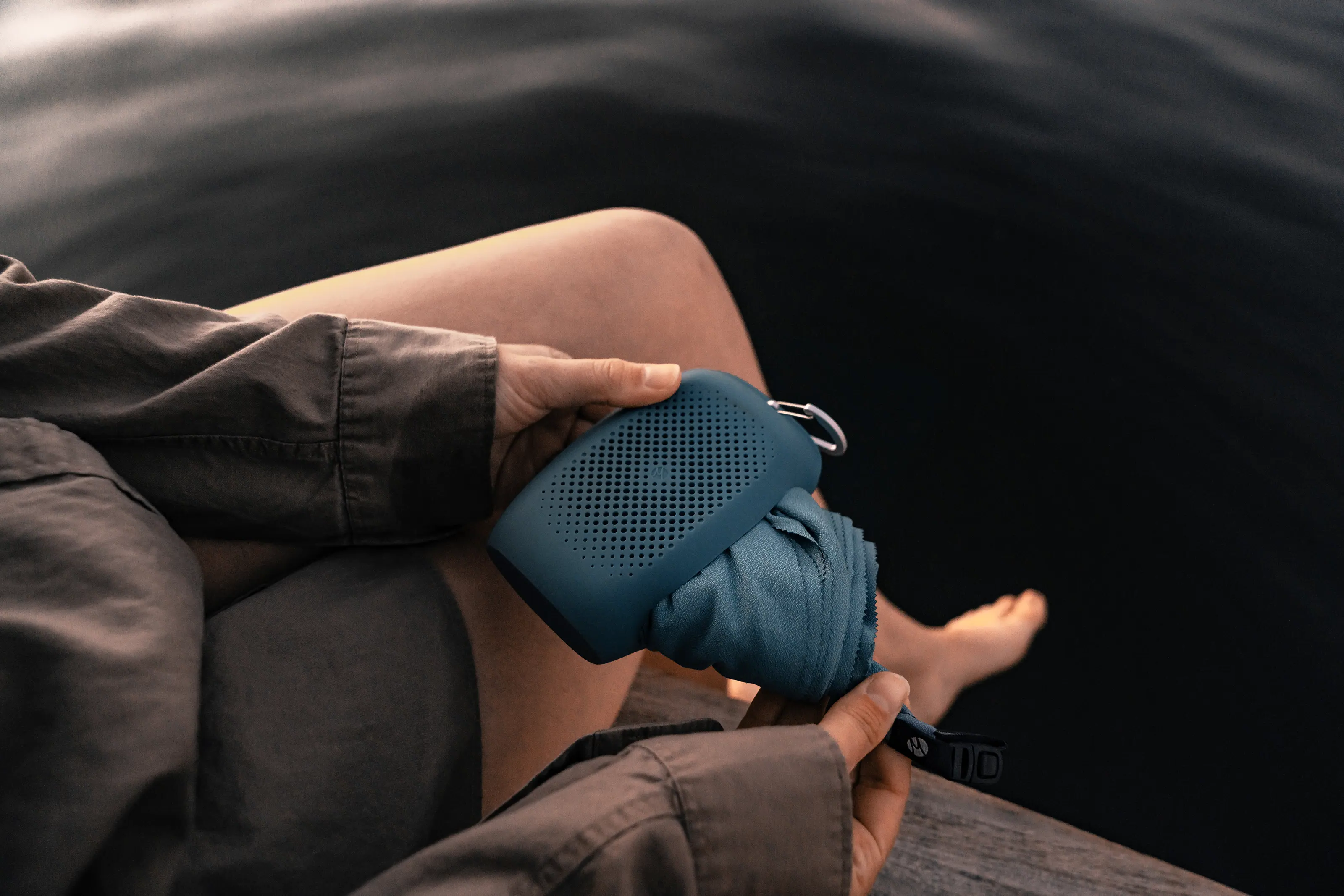 Eine Person mit einem tragbaren Lautsprecher und einem Handtuch in der Hand sitzt am Wasser.