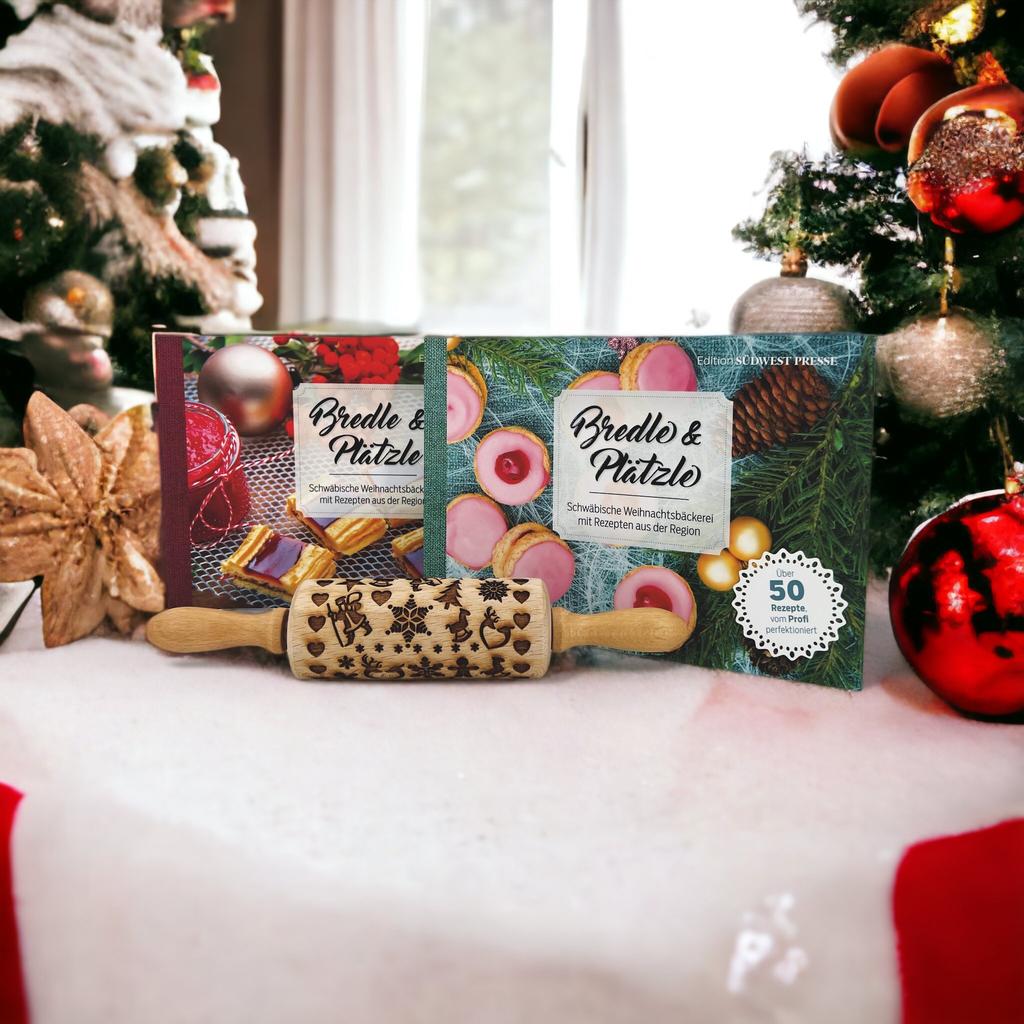 Eine Weihnachtsszene zeigt ein „Set Bredle & Plätzle mit Keksroller“ vor einem Weihnachtsbaum, mit zwei Keksdosen und einem Keksroller mit festlicher Gravur. Der Vordergrund ist mit Ornamenten geschmückt, die eine gemütliche Backenatmosphäre schaffen.