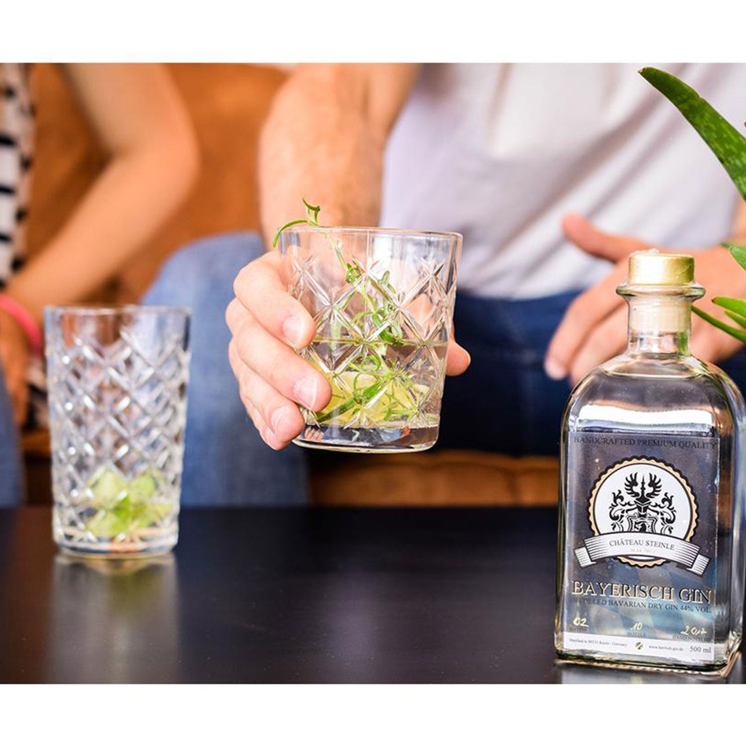 Eine Person hält ein Glas mit einem klaren Getränk, Limette und Bayerischer Gin neben einer Flasche auf dem schwarzen Tisch. Ein weiteres Glas und zwei Personen sind im Hintergrund teilweise sichtbar. Sie sitzen auf einem Sofa und genießen die reichhaltigen Aromen von Château Steinle.