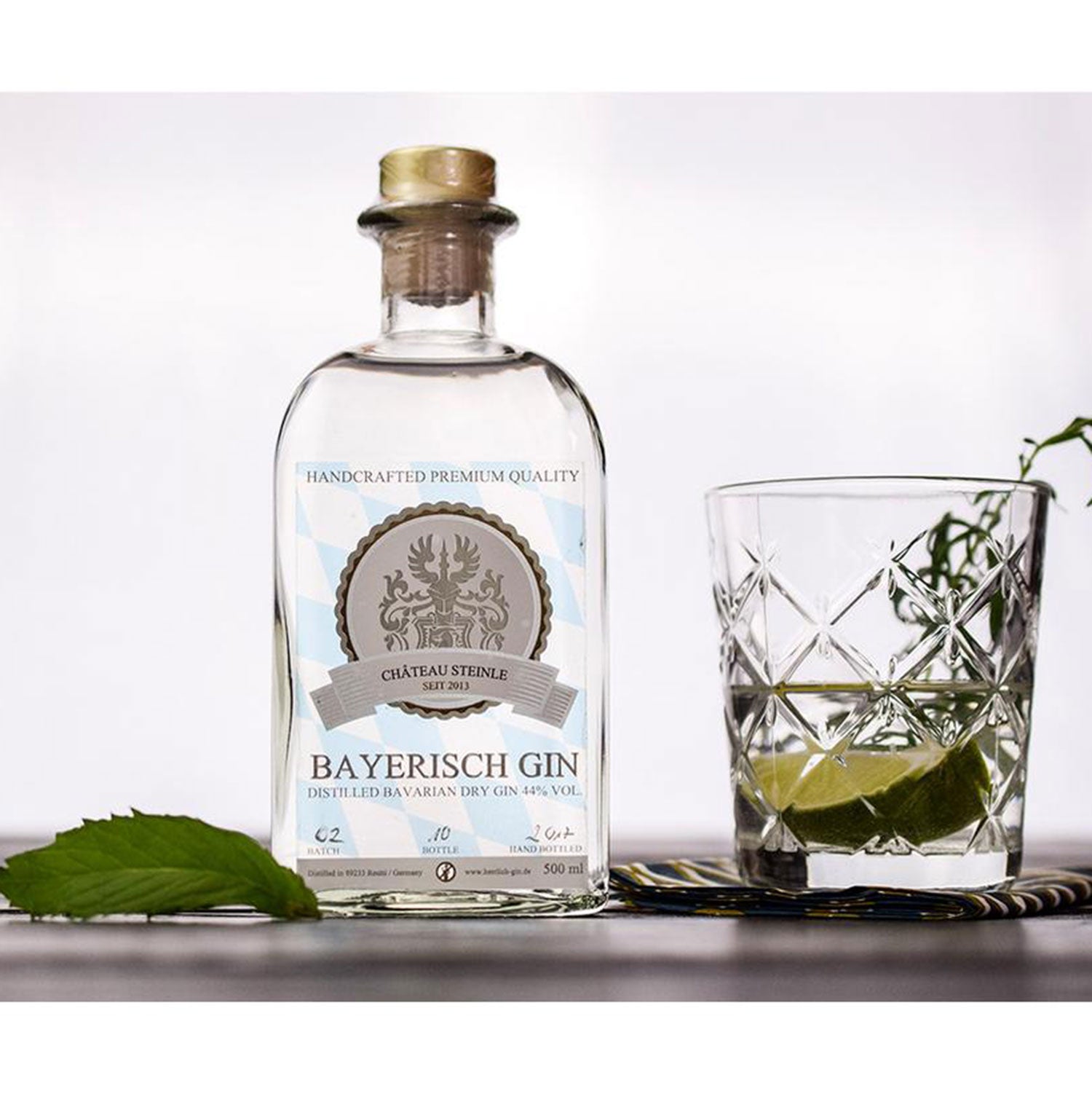 Eine Flasche Bayerischer Gin mit Korkverschluss steht auf einem weißen Untergrund neben einem strukturierten Glas, gefüllt mit Eiswürfeln, Limettenspalten und Botanicals, begleitet von einem grünen Blatt. Das Etikett zeigt ein Wappen und die Aufschrift „Château Steinle“.