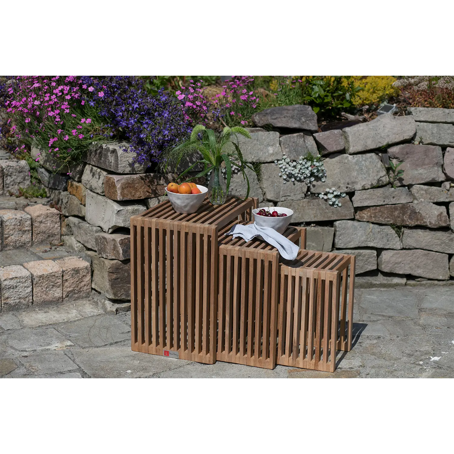 Drei Beistelltische in unterschiedlichen Höhen schaffen eine raffinierte Atmosphäre im Freien: Auf einem stehen eine Pflanze und eine Schale mit Orangen, auf einem anderen sind Kirschen auf einem Tuch ausgestellt, alles vor der Kulisse von Steinstufen und leuchtend violetten Blumen.