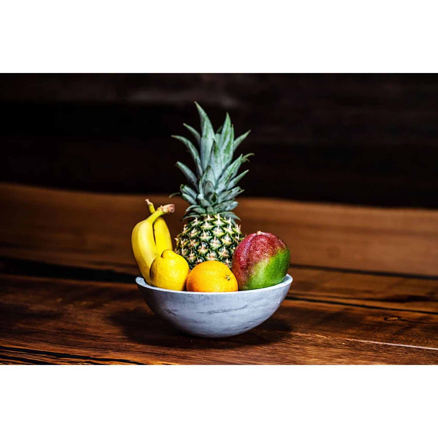 Die „Große Betonschale rund“ mit ihrem handgefertigten, ästhetischen Design steht auf dem Holztisch und enthält Ananas, Bananen, Mango und Orangen. Der dunkle Hintergrund hebt die leuchtenden Farben der Früchte hervor.