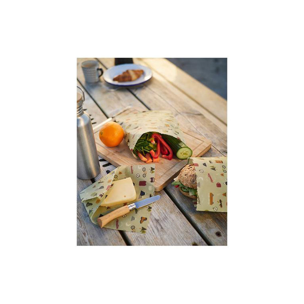 Ein Bio-Bienenwachstuch, ideal zum Frischhalten von Lebensmitteln, umhüllt Gemüse und Käse auf einem hölzernen Picknicktisch. Daneben vervollständigen eine Metallwasserflasche und ein Croissantteller dieses charmante, plastikfreie Outdoor-Set bei Tageslicht.