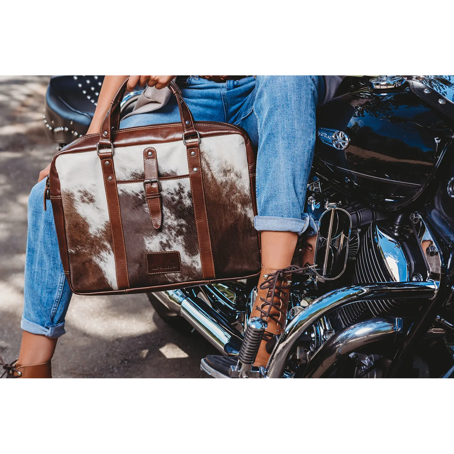 Eine Person sitzt auf einem Motorrad, trägt Bluejeans und braune Schnürstiefel und hält eine einzigartige Aktentasche von LACKL in der Hand. Das Motorrad glänzt mit glänzenden Chromteilen vor einer Außenkulisse.
