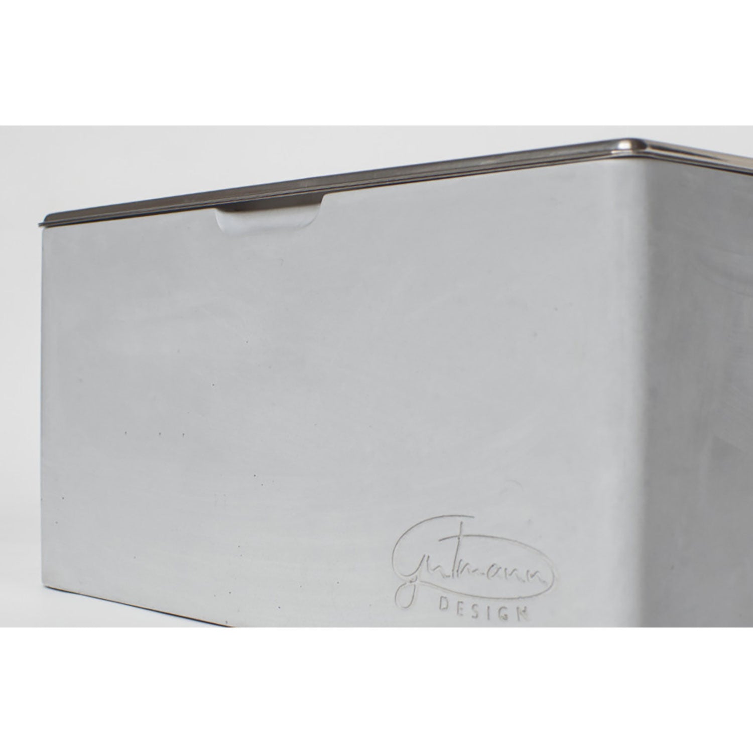Nahaufnahme eines rechteckigen Brotkastens aus Metall mit Edelstahleinsatz und -deckel, glattem hellgrauem Finish und dezentem „Gutmann Design“-Logo an der Seite. Der metallische Glanz des Deckels verleiht ihm einen modernen Touch und erinnert an zeitgenössische Beton-Elemente.