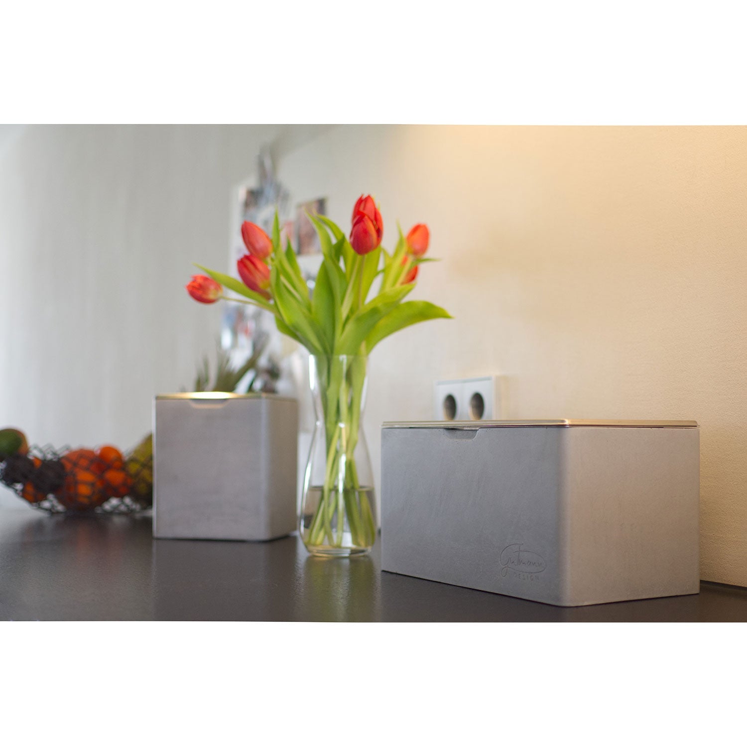 Eine moderne Thekenvitrine präsentiert eine Glasvase mit roten Tulpen, ein elegantes Metallgefäß und einen schicken Brotkasten mit Edelstahleinsatz und -deckel mit Firmenlogo. Ein Obstkorb mit Bananen und Orangen setzt einen farbenfrohen Akzent vor dem minimalistisch-stilvollen Betonhintergrund.