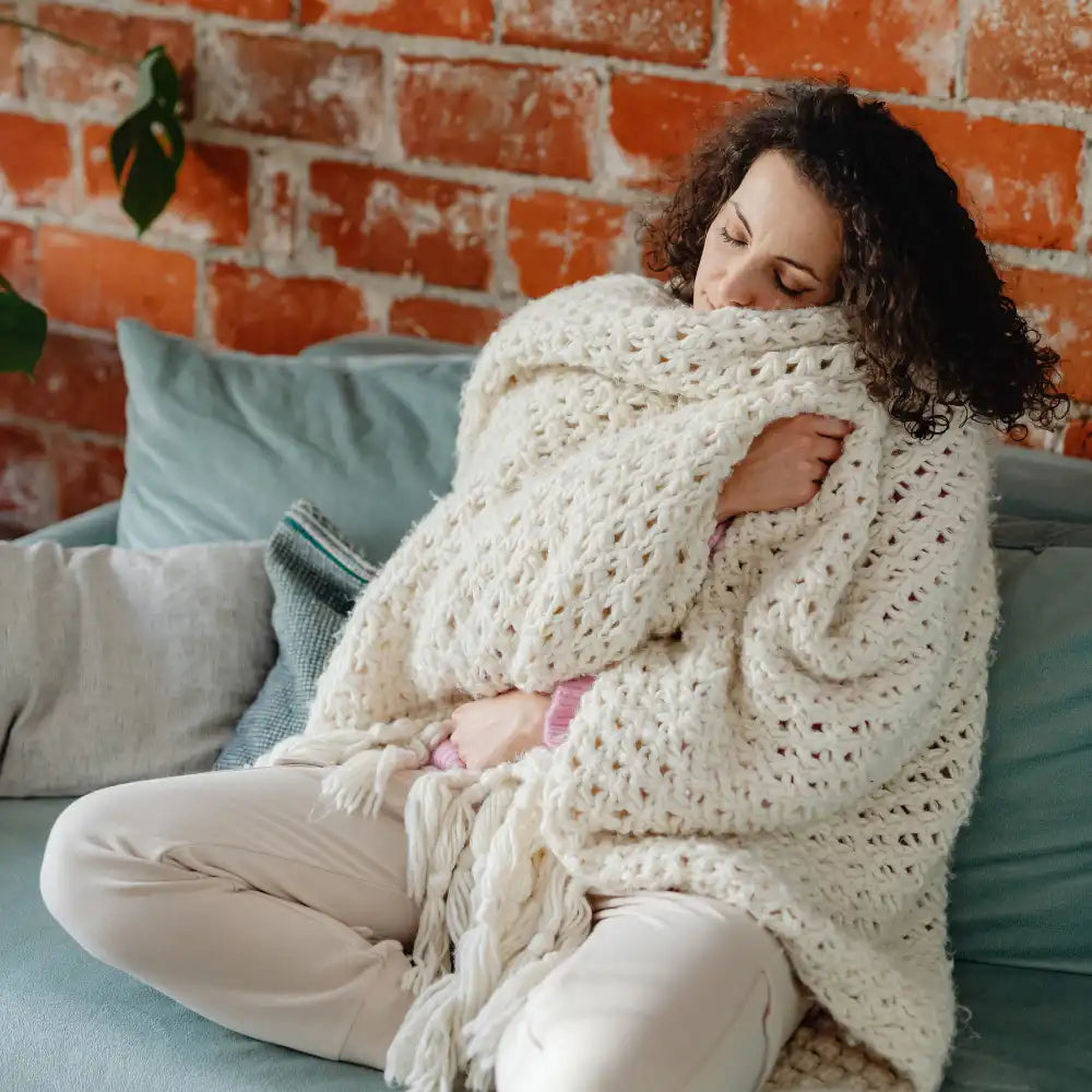 Eine Frau, eingewickelt in eine dicke Strickdecke, sitzt mit geschlossenen Augen auf einer Couch und macht es sich gemütlich.