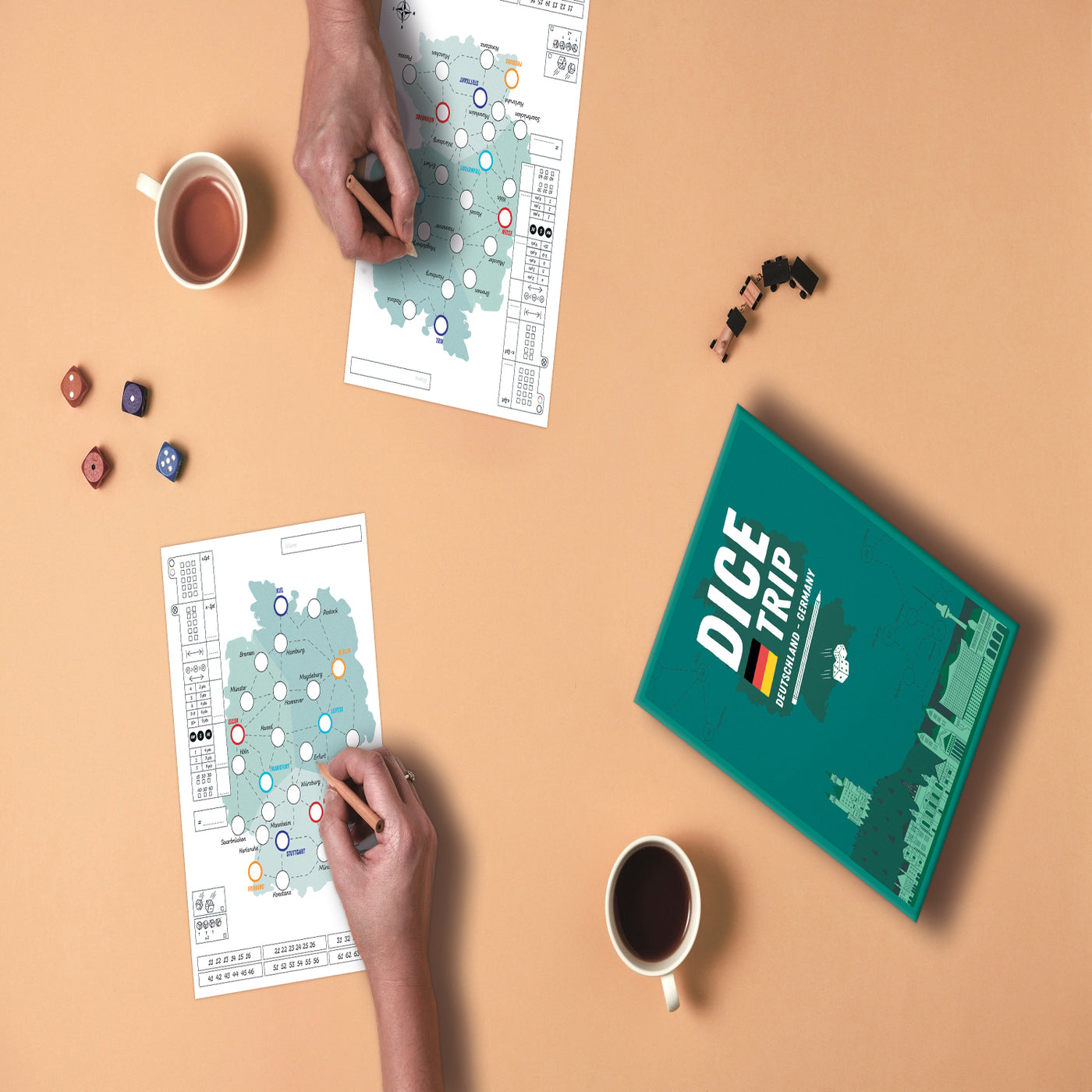 Zwei Personen spielen Dice Trip Deutschland, ein Würfel- und Schreibspiel, mit Würfeln, Spielmarken und Kaffee auf einem hellbraunen Tisch.