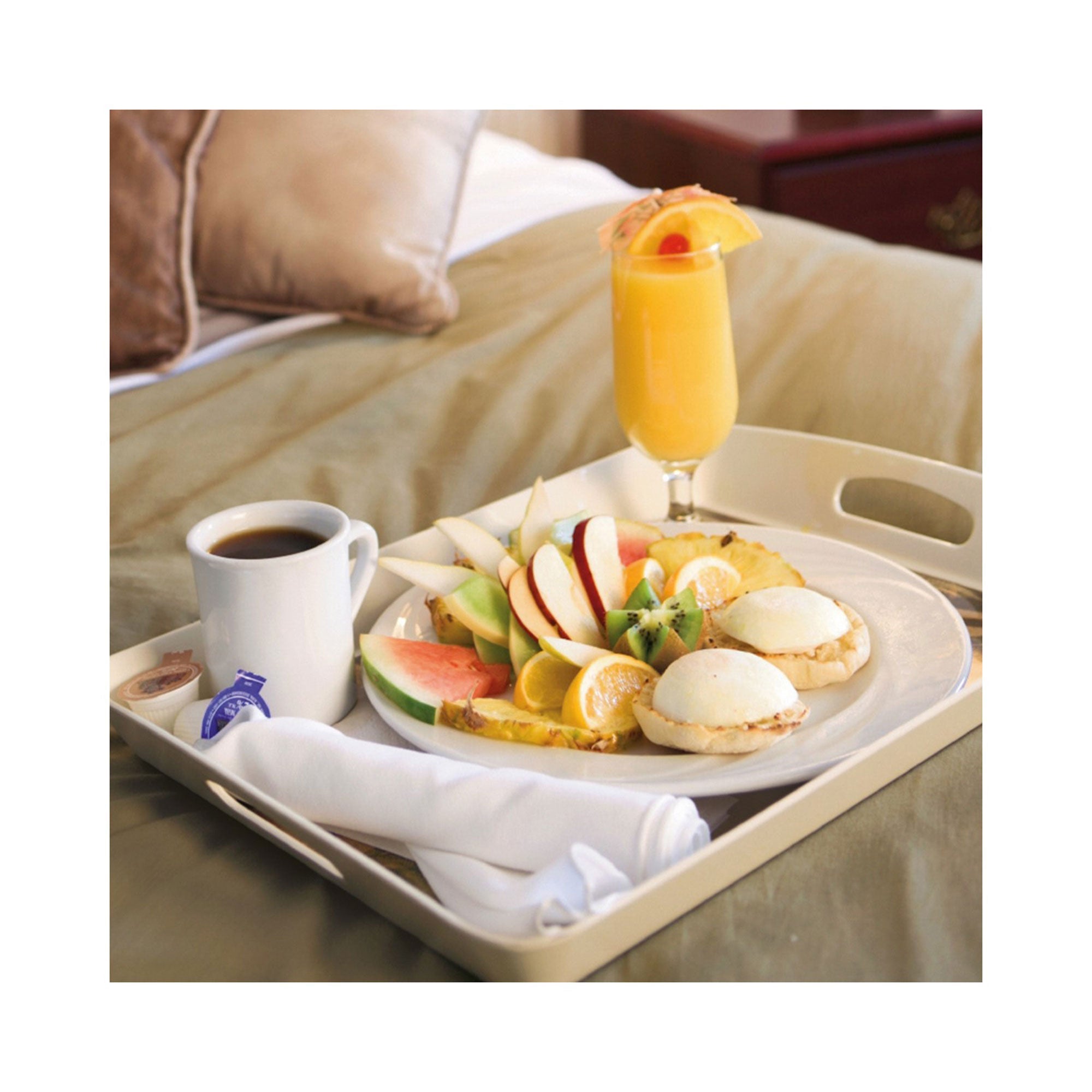 Frühstück im Bett: Ein luxuriöses Tablett mit geschnittenem Obst, aromatisierten englischen Muffins, Kaffee und Orangensaft. Dieser köstliche Morgengenuss fühlt sich an wie eine Auszeit mit der Erlebnis-Box 3 Tage Du & Ich und sorgt für Entspannung zwischen den beige-braunen Kissen.