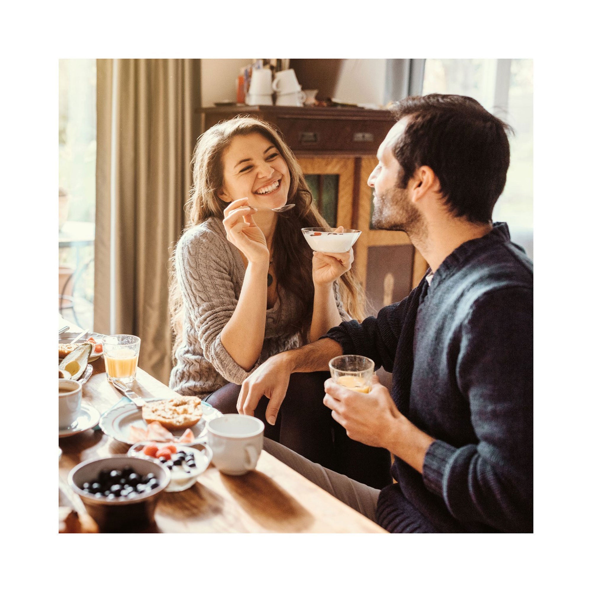 Eine lächelnde Frau und ein Mann genießen ein gemütliches Frühstück, perfekt für einen Kurzurlaub, an einem Holztisch. Sie hält Joghurt in der Hand, er nippt an Saft. Getränke, Pfannkuchen und Obst runden die Szene ab, während natürliches Licht den Raum wie eine warme Erlebnis-Box „Endlich Urlaub“ erfüllt.