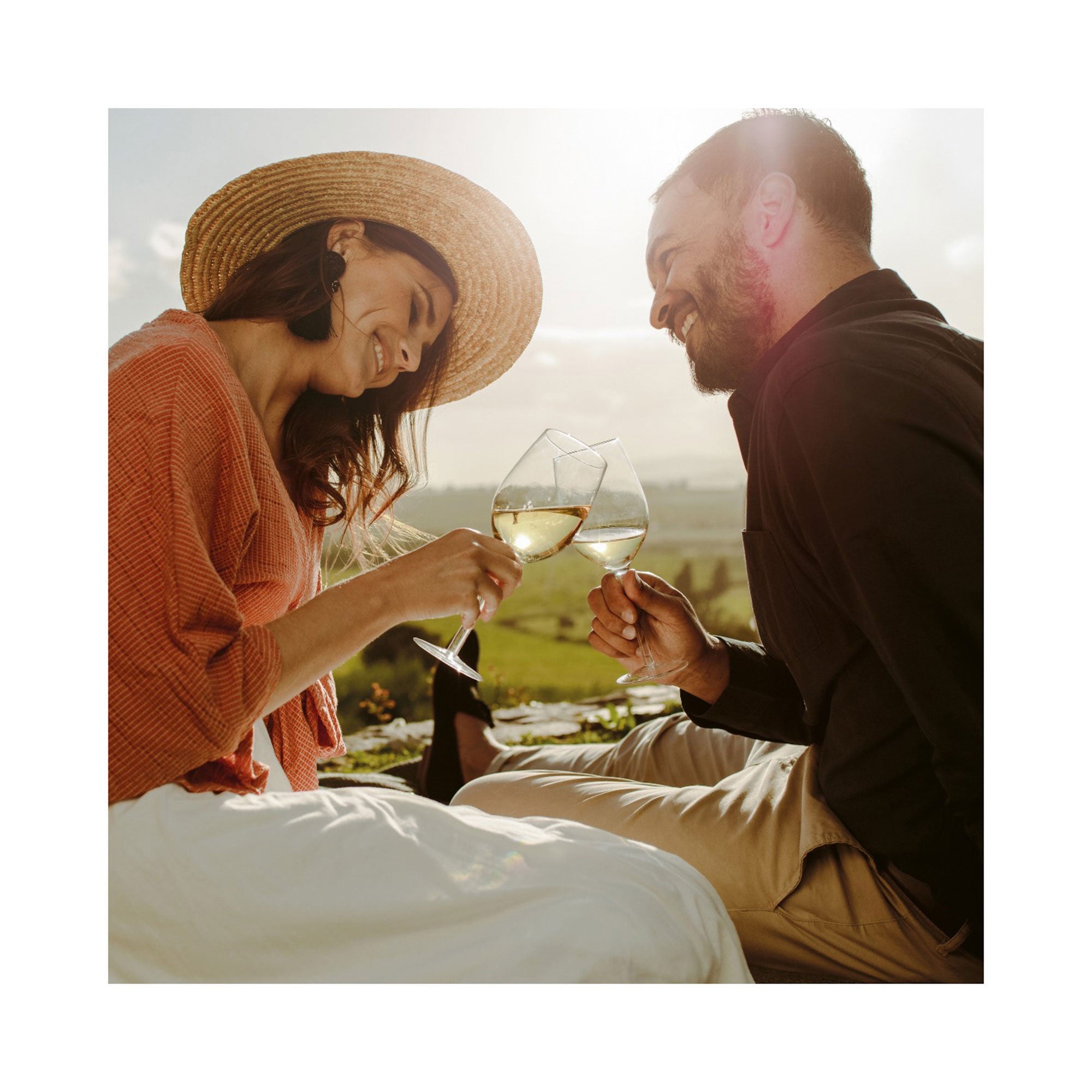 Ein Paar genießt einen Kurzurlaub im Freien mit der Erlebnis-Box „Endlich Urlaub“ und stößt mit Weißwein an. Die Frau mit Strohhut und orangefarbenem Oberteil und der Mann in Schwarz lächeln sich vor der malerischen Kulisse an – ein perfektes Bild der Entspannung.