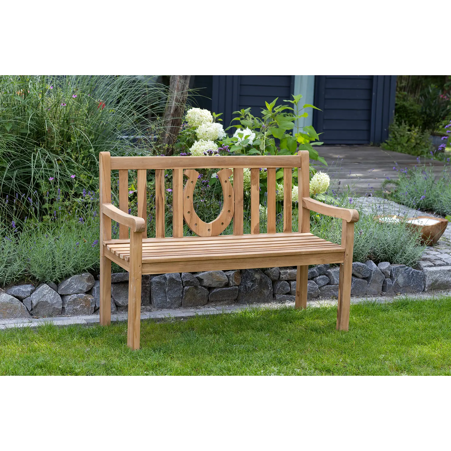 Die Teak-Gartenbank Hufeisen mit ihrer dekorativen Rückenlehne mit Hufeisenmotiv steht auf einem üppigen Rasen, umgeben von steingesäumten Gärten voller vielfältiger Pflanzen und Blumen und lädt zum Entspannen inmitten der Natur ein.