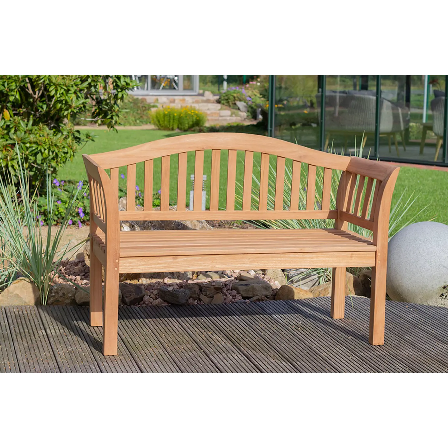 Eine Teak-Gartenbank Wave mit geschwungener Rückenlehne thront elegant auf einem Holzdeck. Sie ist von viel Grün umgeben, und ein großer runder Stein verleiht ihr Charme. Im Hintergrund wertet ein Glasgebäude den üppigen Außenbereich auf.