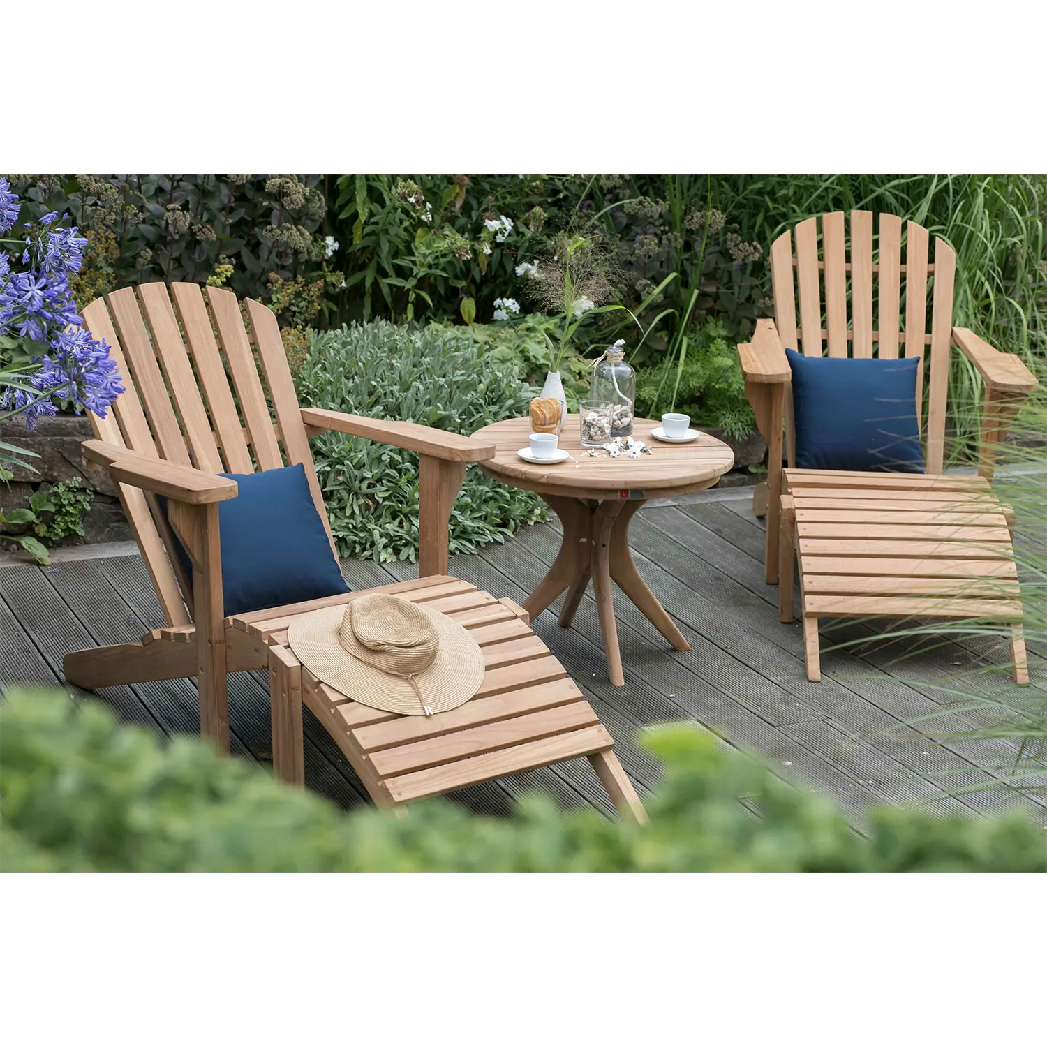 Zwei Gartenliegen aus Teakholz mit blauen Kissen stehen auf einer Terrasse, gegenüber einem runden Tisch mit Teeservice. Auf einer Liege ruht ein Strohhut, während üppige grüne Pflanzen dieses einladende Outdoor-Möbelstück umgeben.