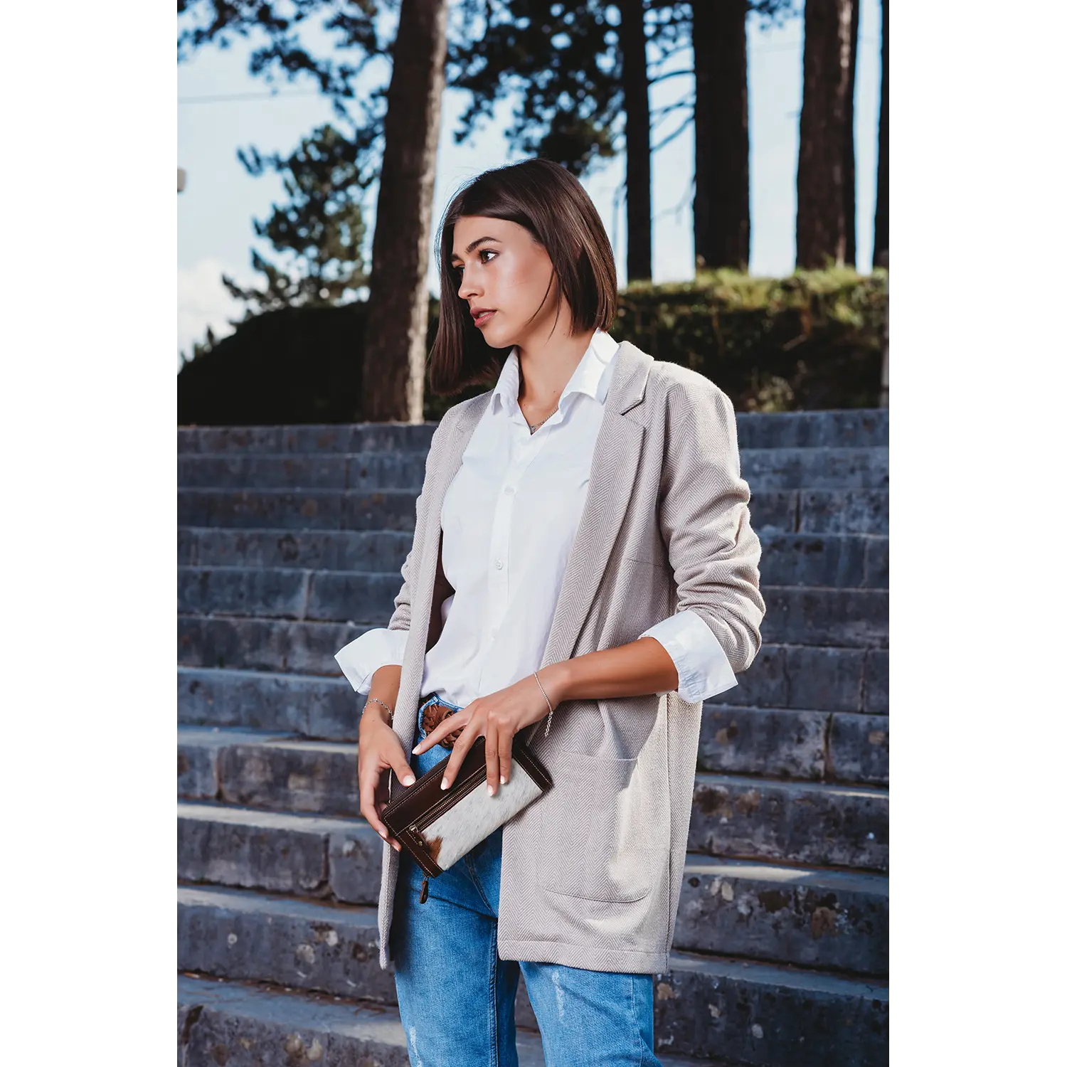 Auf Steinstufen hält eine kurzhaarige Frau in weißem Hemd, hellgrauem Blazer und Bluejeans einen Damengeldbeutel GSPUSI mit RFID-Schutz in der Hand. Im Hintergrund sind üppige Bäume zu sehen.