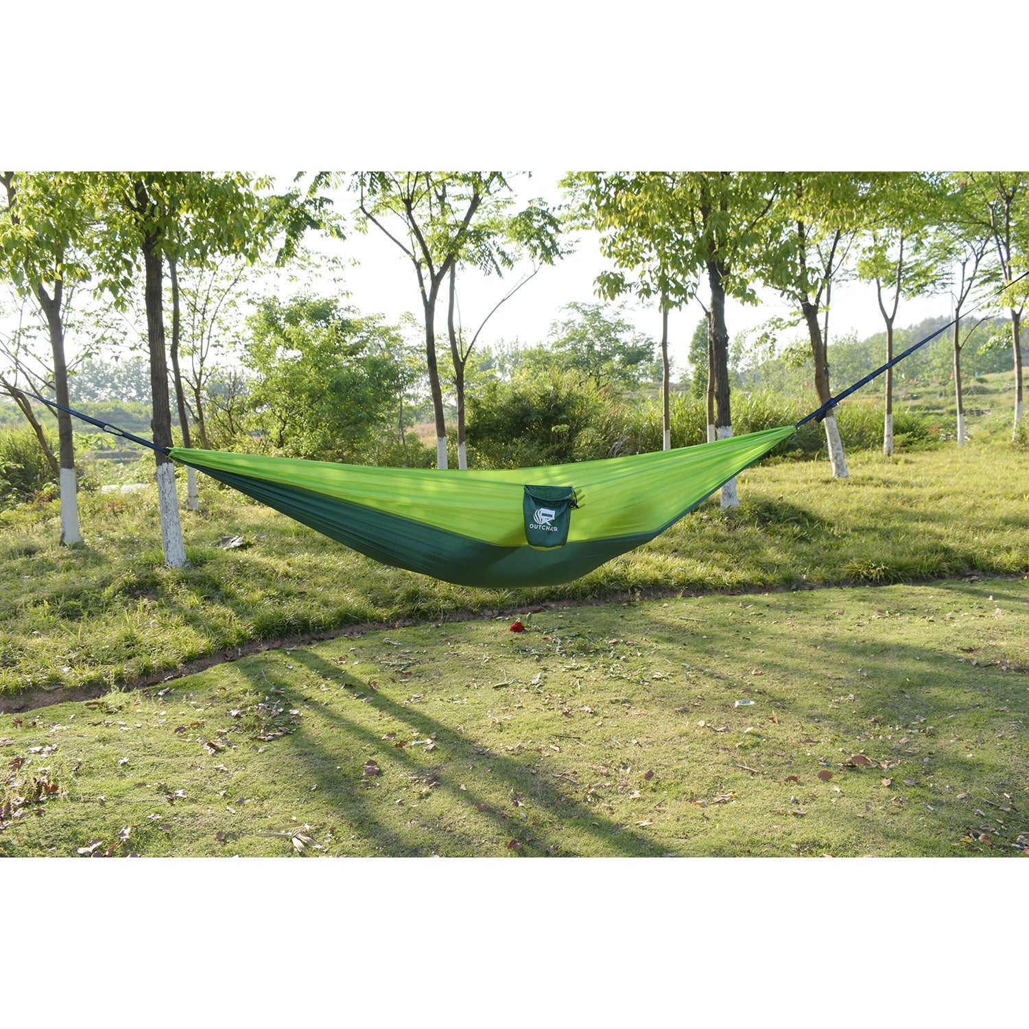 Eine superleichte Hängematte in Grün hängt zwischen zwei Bäumen in einem sonnenbeschienenen Park. Sie ist auf Langlebigkeit ausgelegt, verfügt über eine kleine Seitentasche und bleibt ungenutzt, während sich lange Schatten über den Boden ziehen.