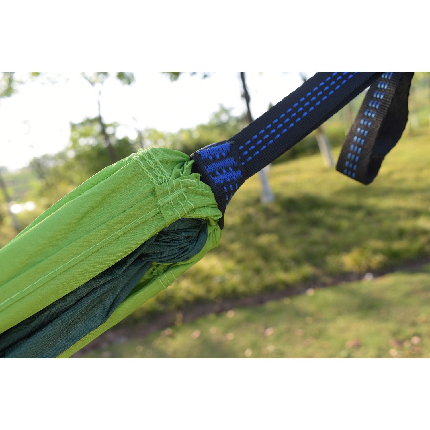 Nahaufnahme eines grünen Gurtes an einer superleichten Hängematte, befestigt an einem blau-schwarzen Seil vor einem verschwommenen Hintergrund aus Gras und Bäumen. Das Bild hebt die strapazierfähigen Nähte und die Stoffstruktur hervor und verkörpert so ihren robusten Charakter für leicht transportierbare Abenteuer.