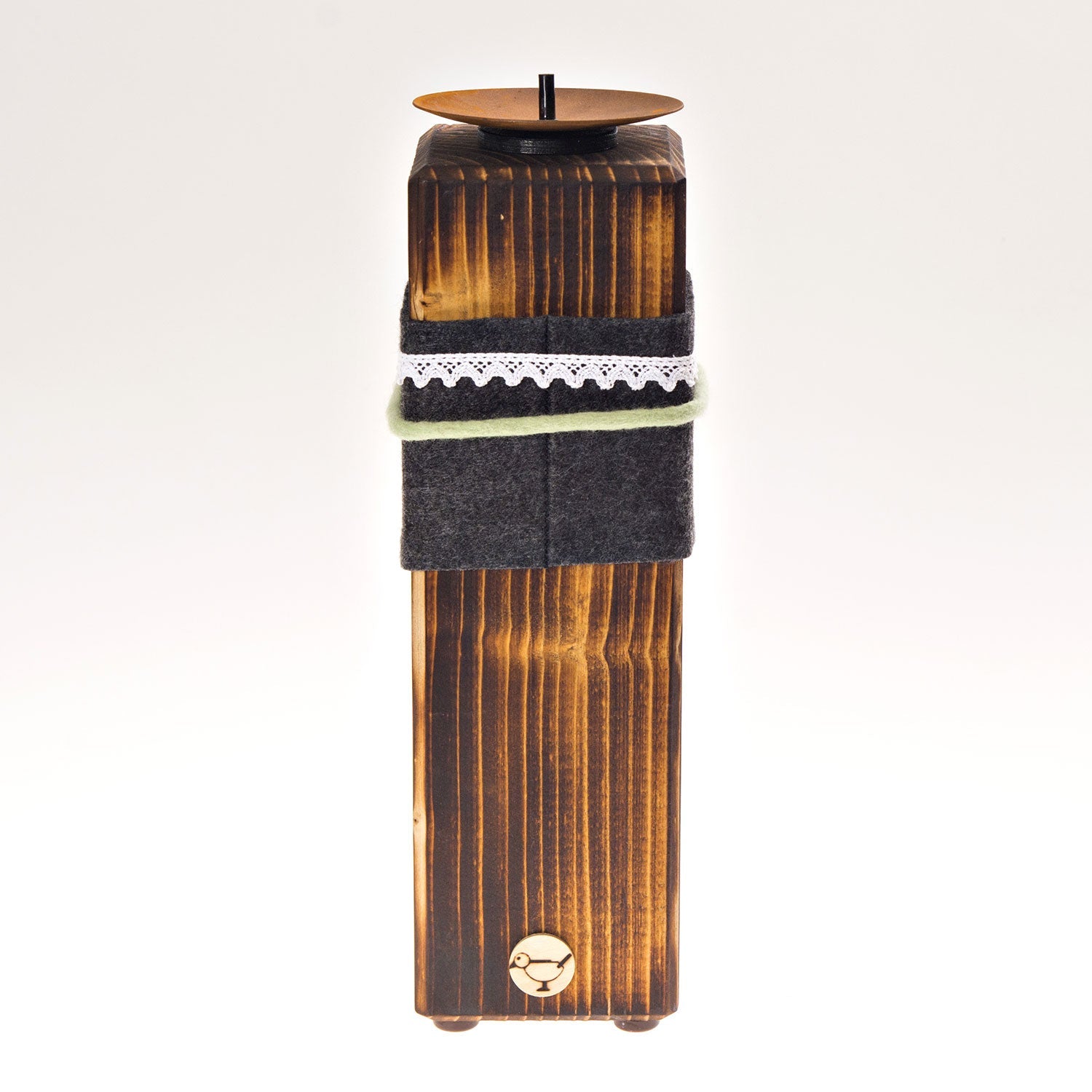 Der „Kerzenständer aus Holz – groß“ ist ein rustikales Holzstück mit einem schwarzen, zylindrischen Kerzenhalter. Er ist aus Massivholz gefertigt, mit grauem Stoff und einem Spitzenband versehen und mit einem kleinen Logo am Fuß versehen. Ideal für die Präsentation von Stumpenkerzen auf dem schlichten weißen Hintergrund.