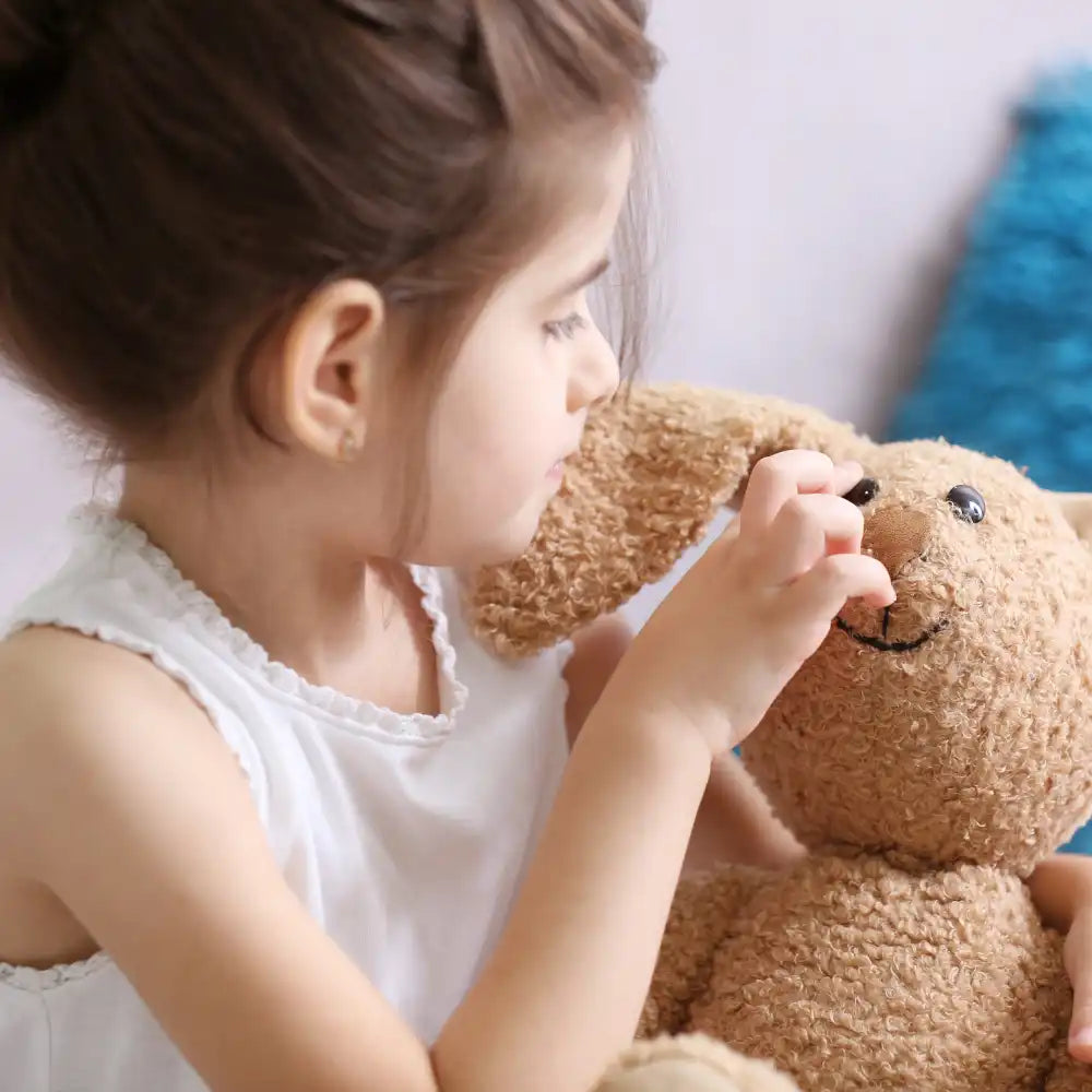 Junges Mädchen in einem weißen Oberteil, das das Gesicht eines braunen Stofftiers berührt.