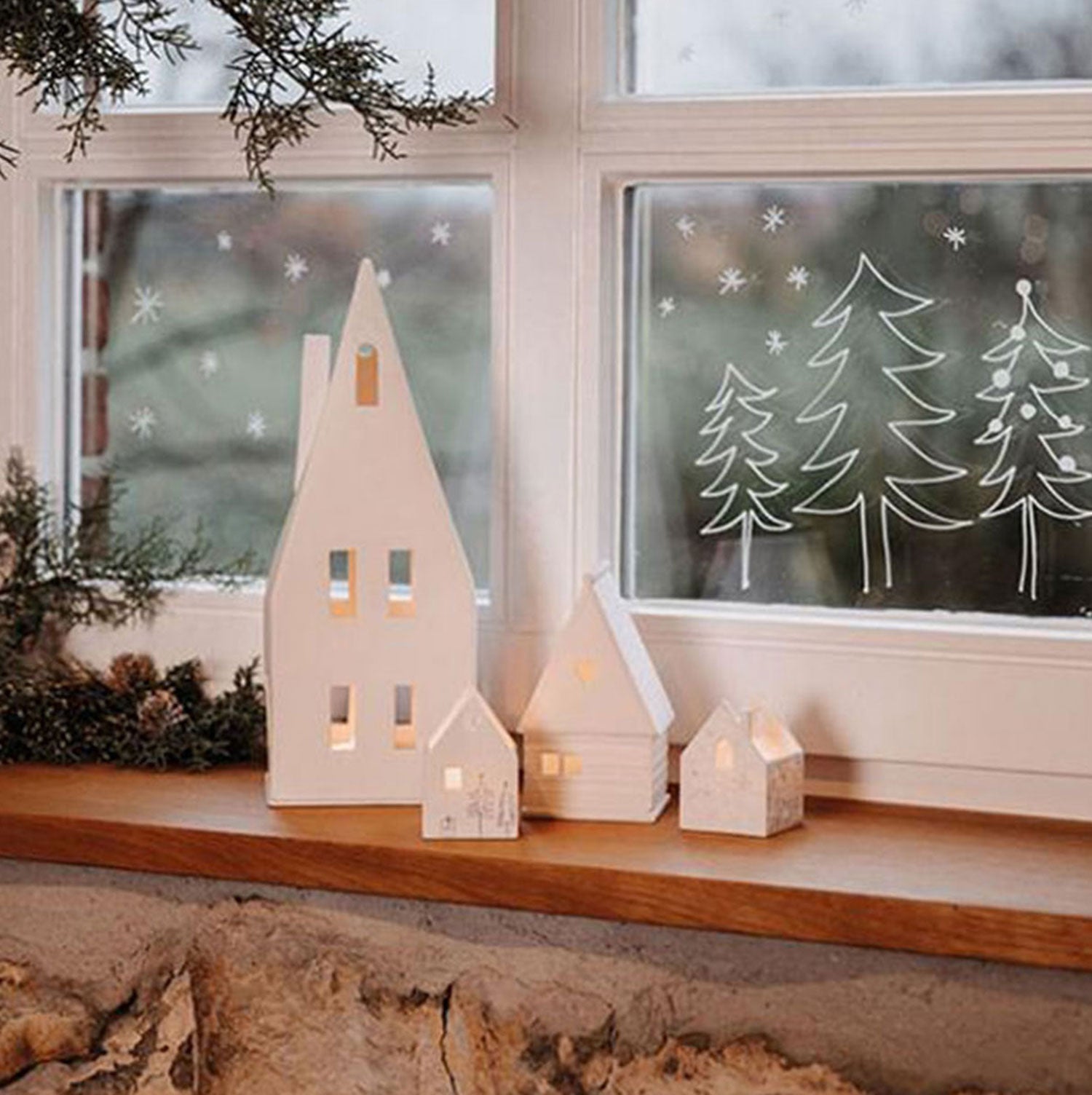 Eine Fensterbank mit kleinen, weißen Laternen „Spitzdach“ von Großes Lichthaus strahlt warmes Licht aus. Das Fenster ist mit weiß bemalten Baum- und Schneeflockenmotiven geschmückt, eingerahmt von immergrünen Zweigen. Unter dem Holzregal befindet sich eine Steinmauer, die eine gemütliche Winterlandschaft mit handgefertigtem Charme schafft.