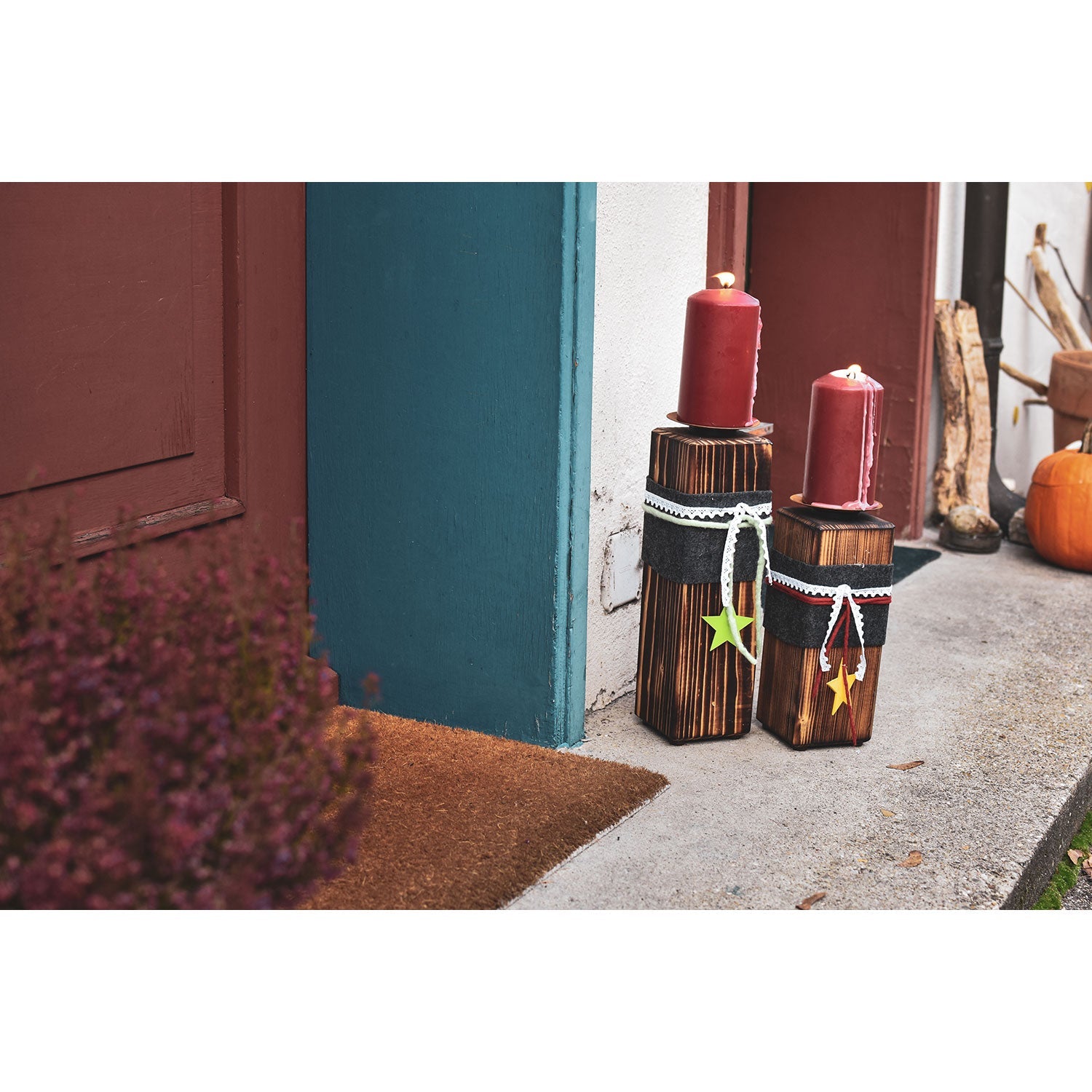 Der große Kerzenständer aus Holz, verziert mit Sternornamenten, steht neben einer blau-braunen Tür auf einer Veranda. Ein Kürbis im Hintergrund sorgt für gemütliche Herbststimmung und ergänzt die eleganten Massivholz-Akzente.