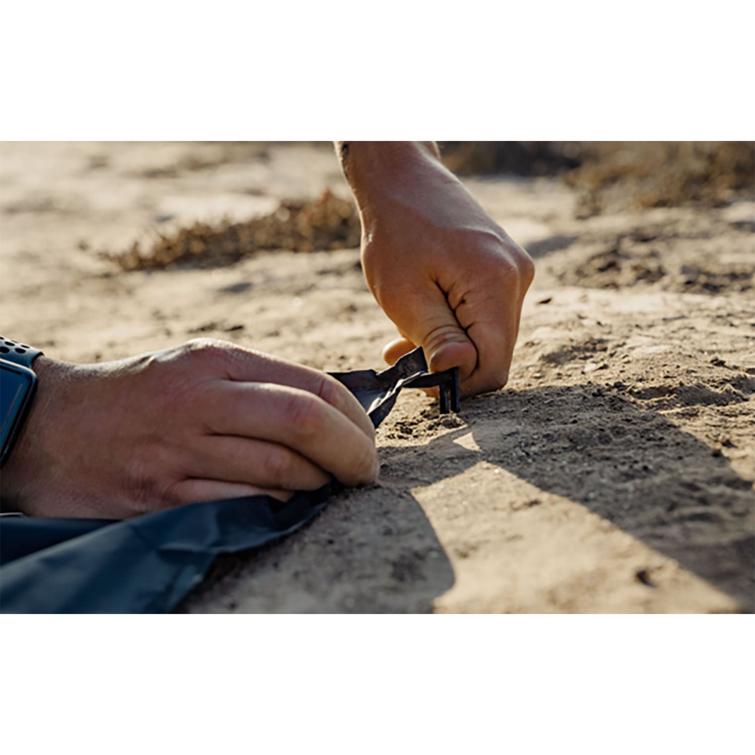 Nahaufnahme von Händen, die einen Zelthering im Sandboden befestigen. Eine Hand hält ein Werkzeug, die andere den Hering. Die Person mit der Uhr stellt sich in trockener Umgebung im Freien neben ihre wasserabweisende Outdoor-Decke mit Befestigungshaken.