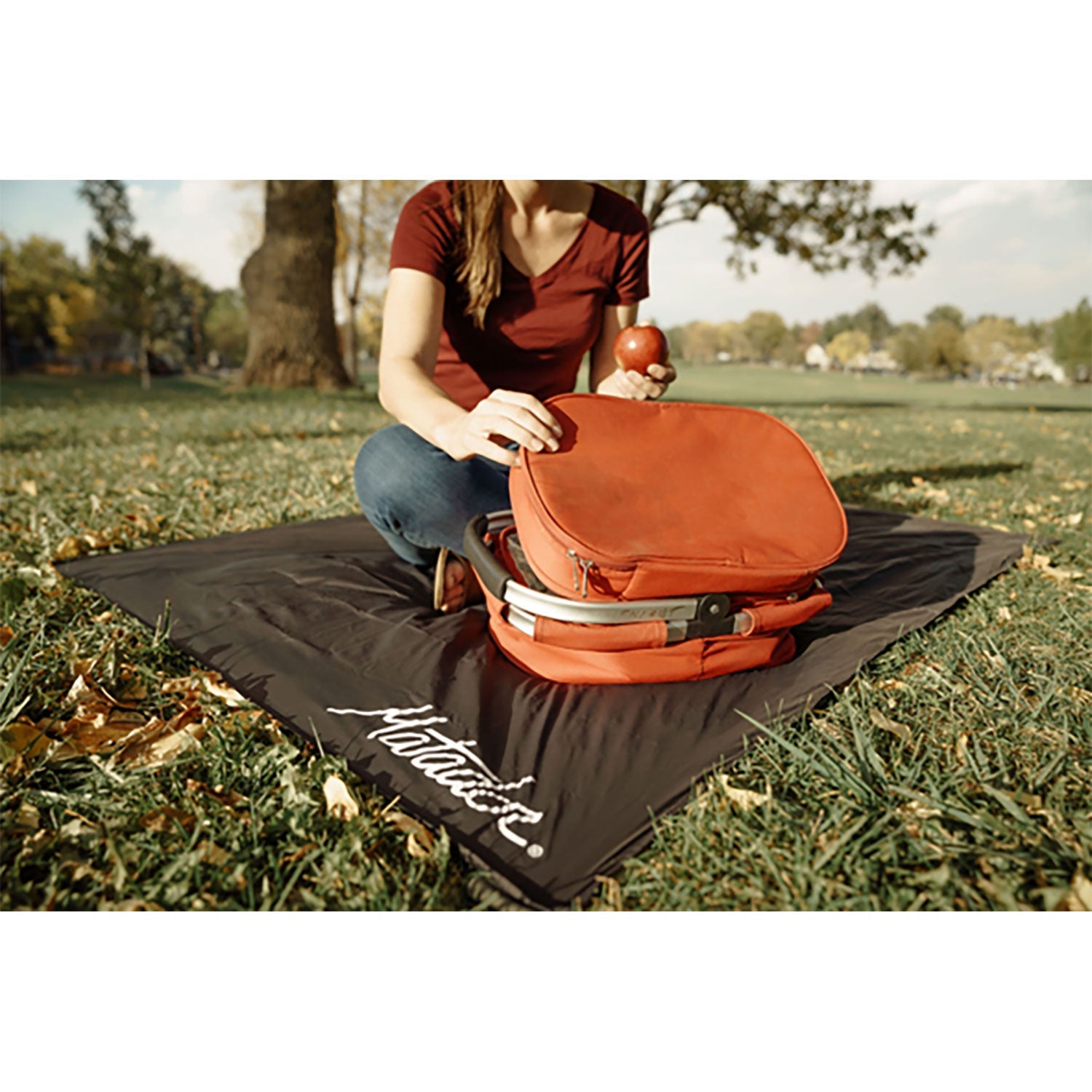 Eine Person in einem roten Hemd und Jeans genießt den Komfort ihrer schwarzen Outdoor-Decke mit Befestigungshaken im Park, hält einen Apfel in der Hand und öffnet einen orangefarbenen Rucksack, umgeben von grünem Gras und Bäumen.