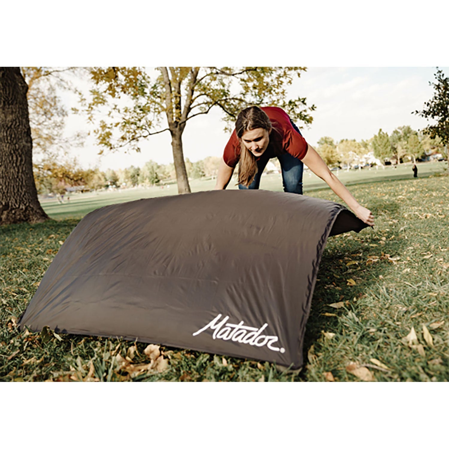 Eine Person breitet eine große, braune Outdoor-Decke mit Befestigungshaken auf einer Wiese aus. Im Hintergrund sind Bäume mit Herbstlaub unter einem bewölkten Himmel zu sehen. Die Person trägt ein kastanienbraunes Hemd und Jeans.