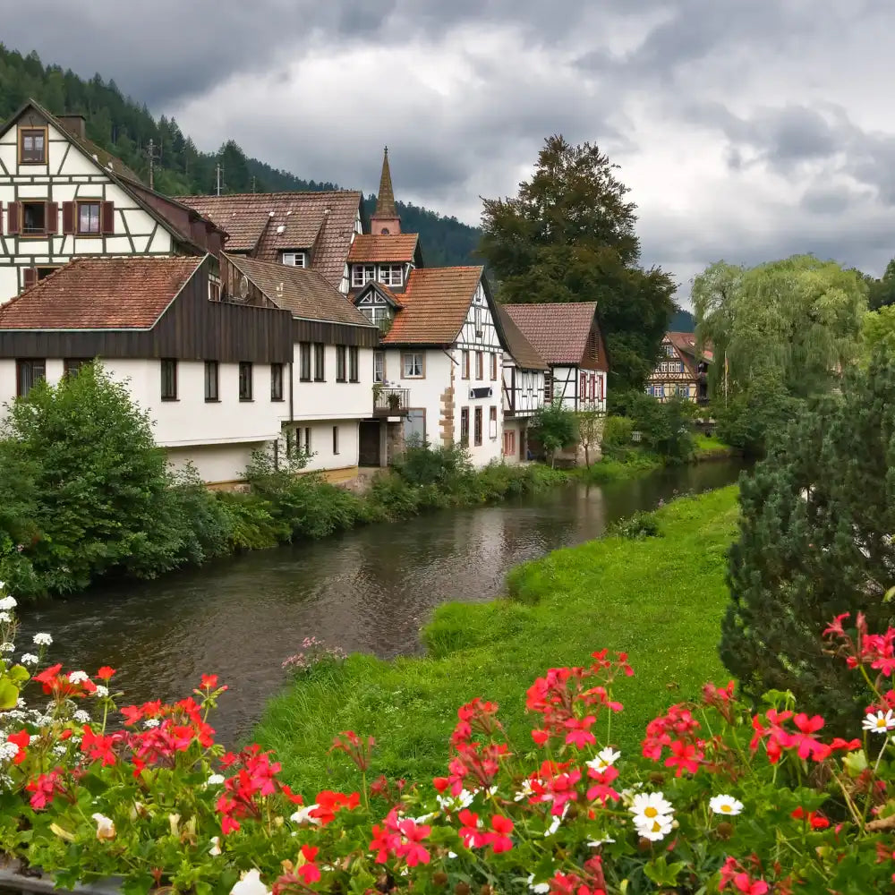 Malerische Dorfhäuser an einem Fluss mit Blumen und Grünpflanzen unter einem wolkenverhangenen Himmel.