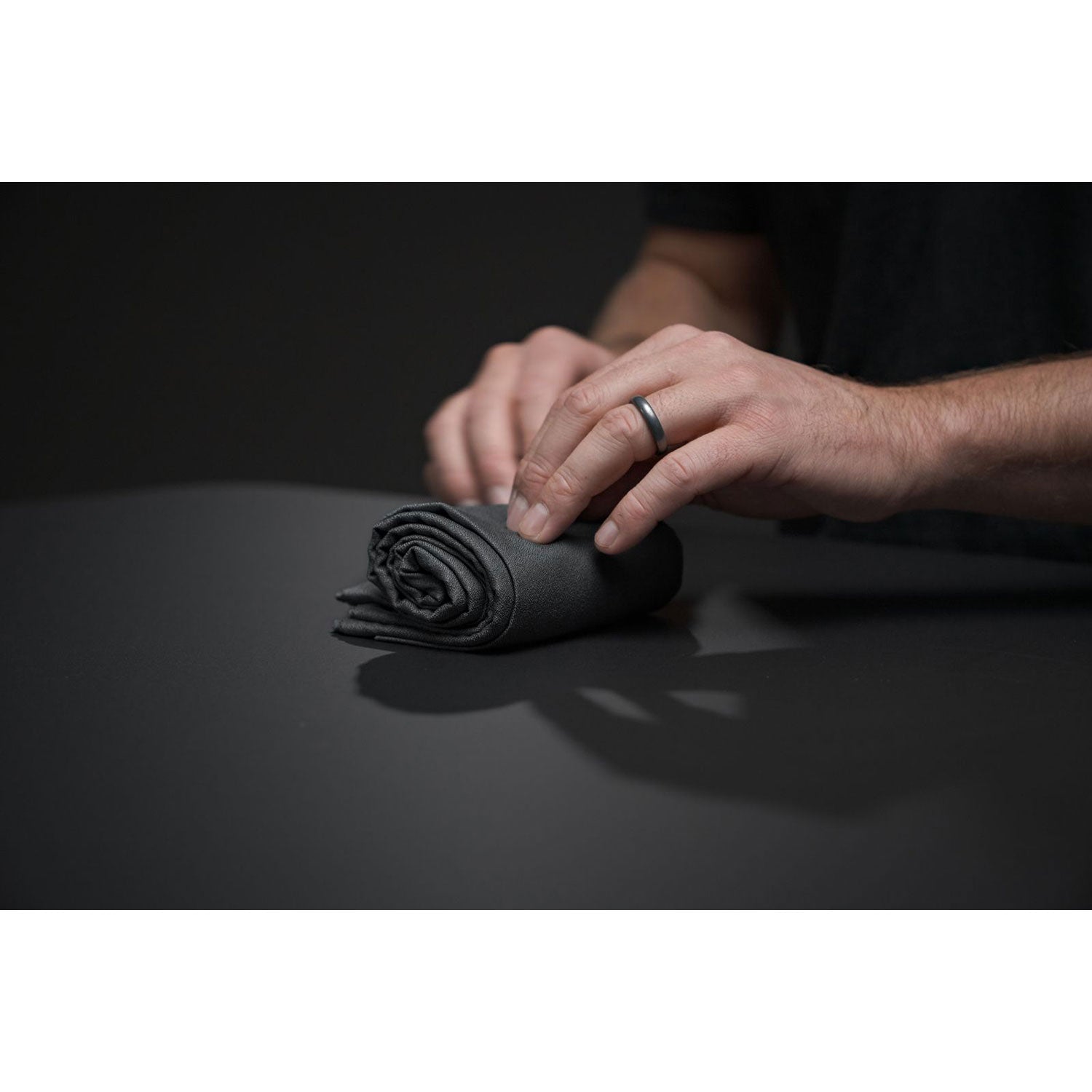 Eine Person mit einem Ring an der linken Hand rollt vor schwach beleuchtetem Hintergrund vorsichtig ein ultraleichtes Nanodry Handtuch mit dunkler, antimikrobieller Beschichtung auf einem schwarzen Tisch.