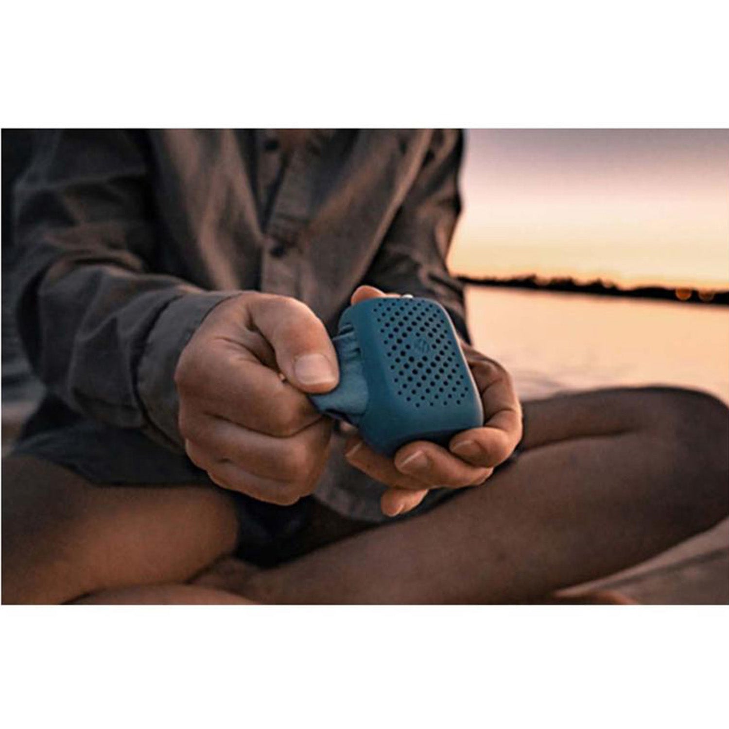 Eine Person sitzt im Schneidersitz im Freien mit einem Matador Nanodry Reisehandtuch und präsentiert es als ideale platzsparende Ausrüstung. Das Reisehandtuch ist das Herzstück vor der Kulisse eines Sonnenuntergangs über dem Wasser und verkörpert perfekt spontanes Abenteuer in lässiger Kleidung.