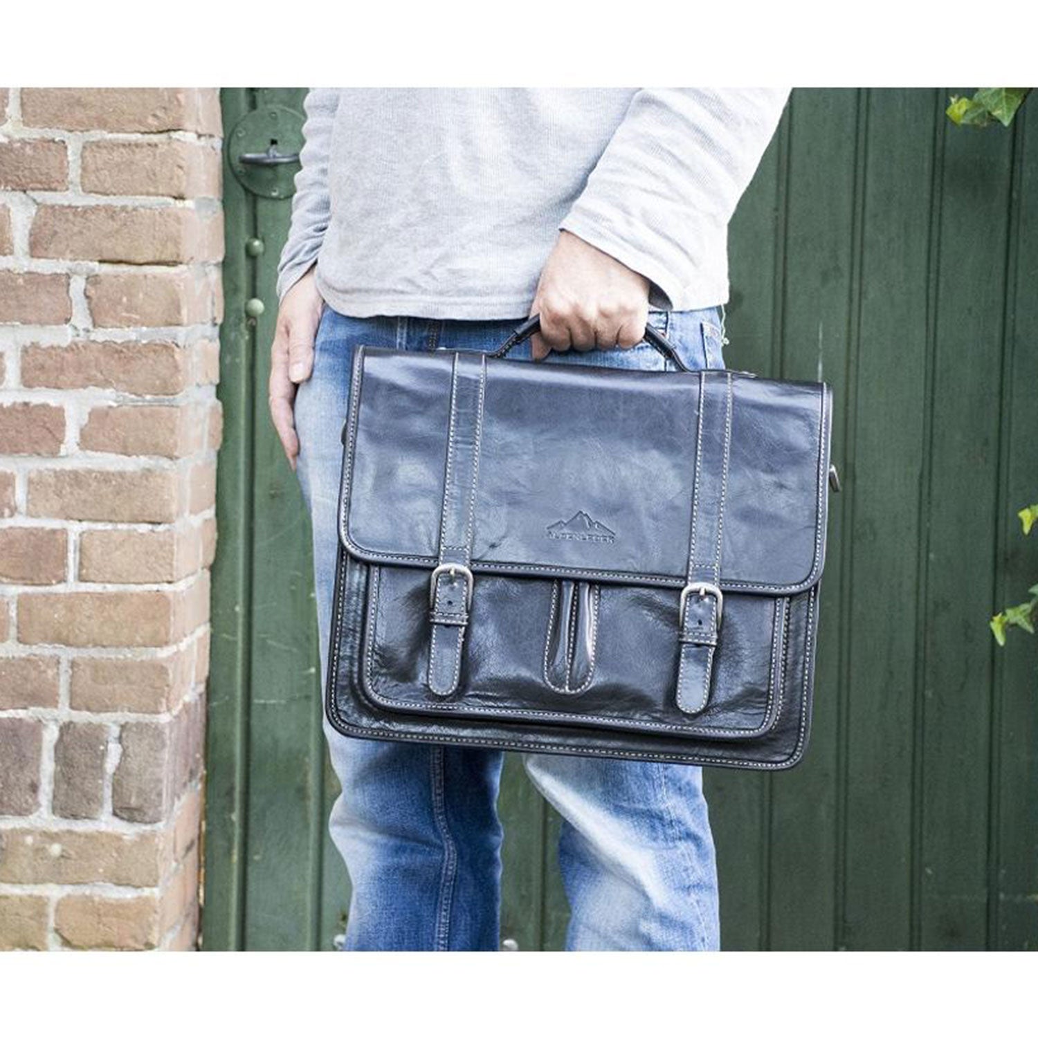 Eine Person in grauem Hemd und Jeans steht vor einer grünen Tür und einer Backsteinmauer. Sie hält eine schwarze Aktentasche OHIO aus Leder mit Schnallen und Tragegriff in der Hand. Die Aktentasche verfügt über ein praktisches Laptopfach.