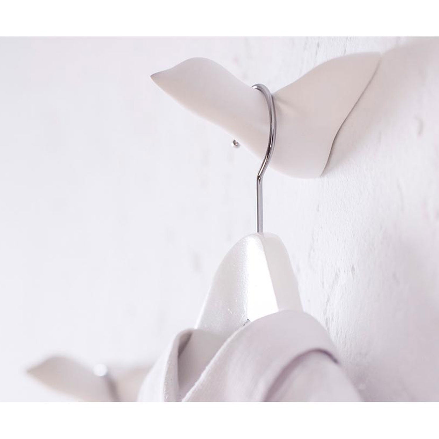 Nahaufnahme eines Wandskulptur-Mini-Vogel-Wandhakens, der einen Metallbügel mit einem weißen Kleidungsstück vor einer weißen Wand hält. Ein weiterer ähnlicher Wandskulptur-Mini-Vogel-Haken ist im Hintergrund teilweise sichtbar.