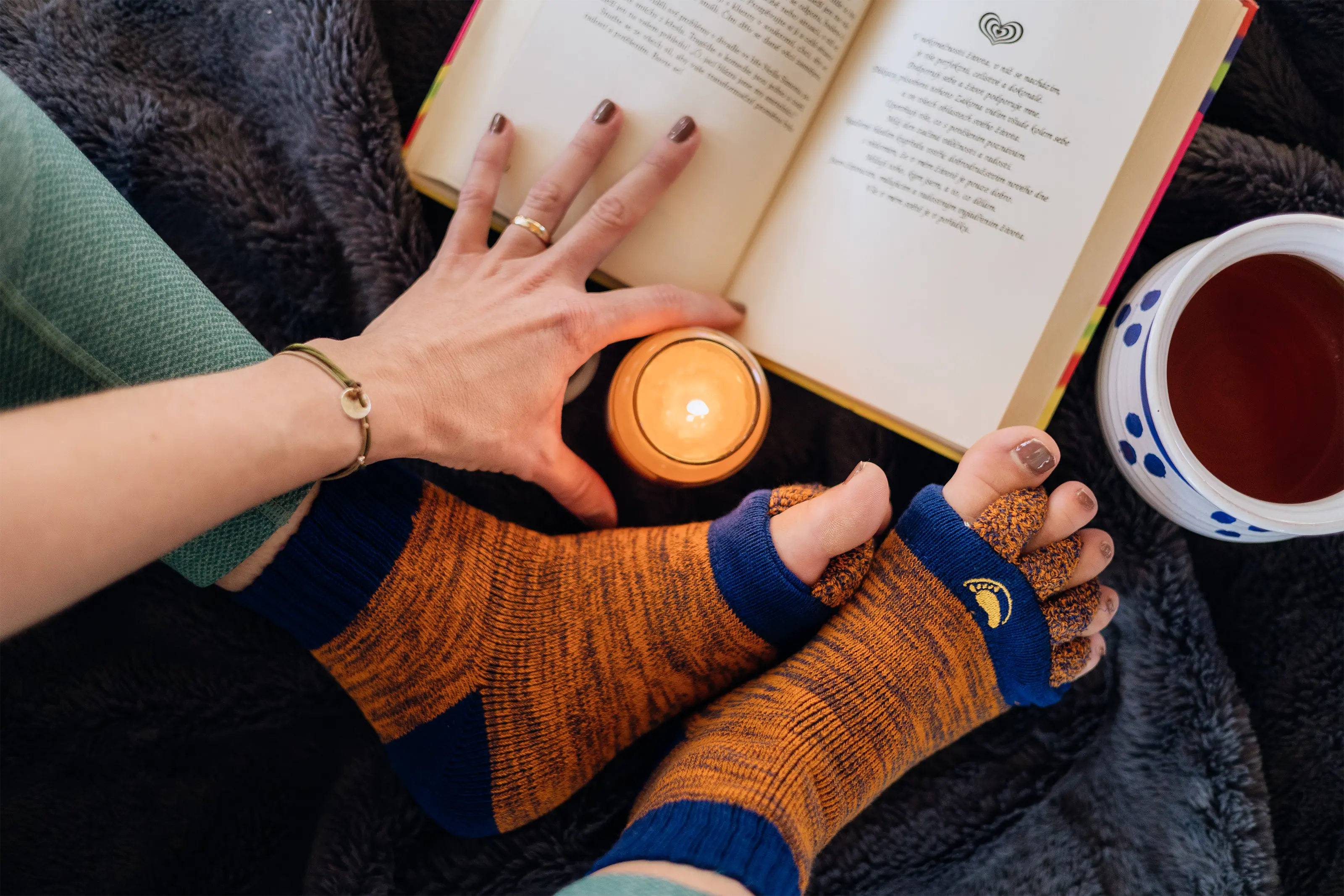 Eine Person, die orangefarbene Zehensocken trägt, liest ein Buch mit Tee und einer brennenden Kerze in der Nähe.