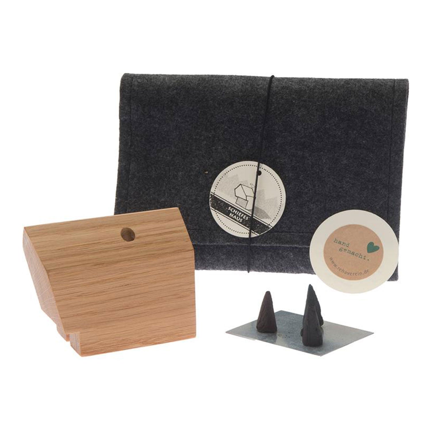 Das Räucherhäuschen „Schiefes Haus“ mit zwei schwarzen Tannenduftkegeln auf einem Metallquadrat wird von einem Filzbeutel mit kreisförmigem Logo begleitet, das die Essenz der Weihnachtstradition einfängt.