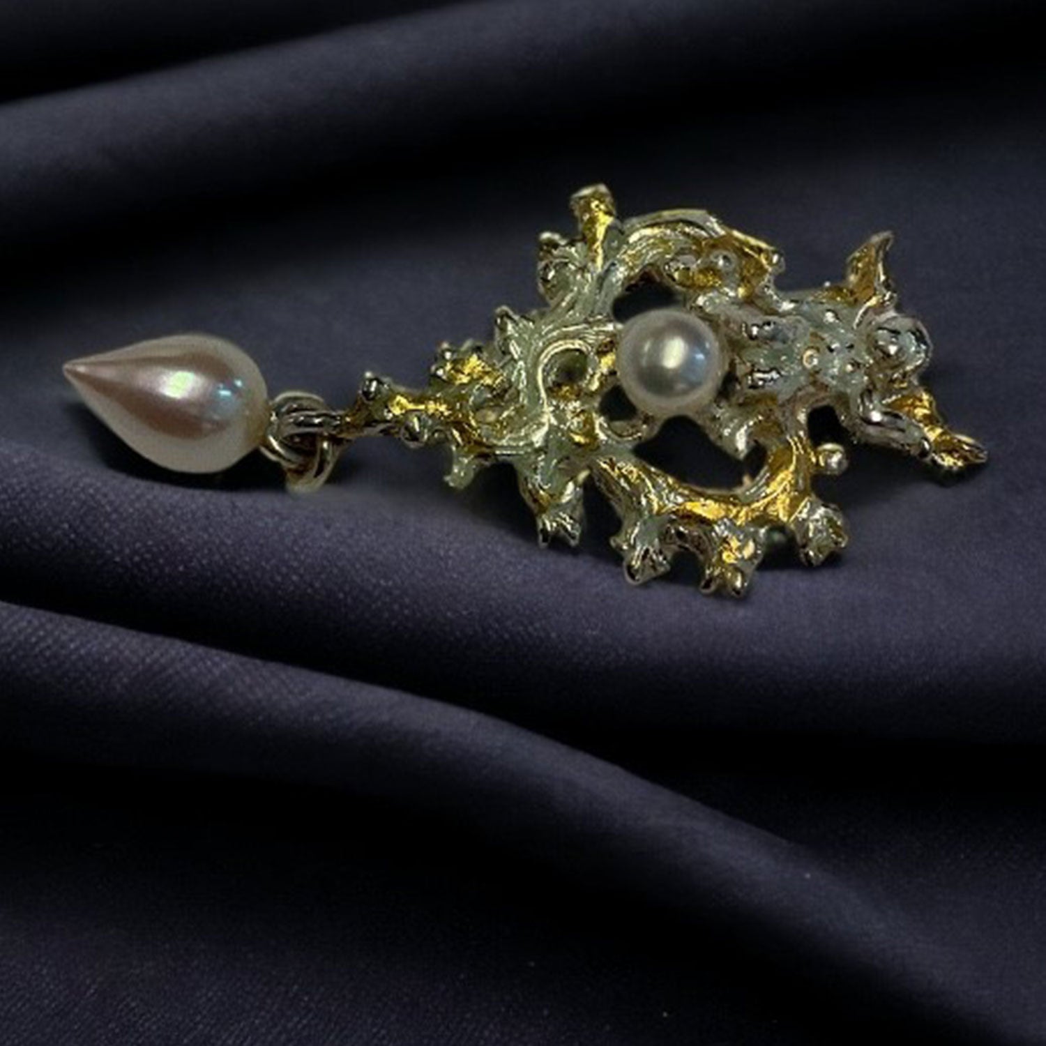 Der Anhänger Sanssoucis Engel ist eine luxuriöse Brosche aus 925/- Sterlingsilber mit einem filigranen Gold- und Silberdesign. Er besticht durch eine zentrale Perle mit wellenförmigem Metallmuster und eine darunterliegende tropfenförmige Perle, die vor dunklem Stoffhintergrund Feingold-artige Eleganz ausstrahlt.