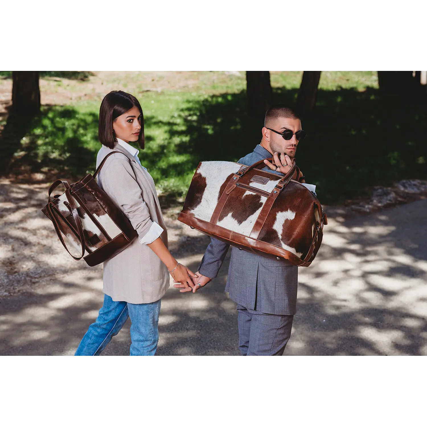 Ein Paar geht Hand in Hand. Die Frau trägt Blazer und Jeans und trägt eine große braun-weiße Alpenleder-Shopper-HALLODRI-Tasche über der Schulter. Der Mann mit Sonnenbrille und Anzug trägt eine passende Kuhfell-Reisetasche. Sie schlendern den sonnenbeschienenen Weg entlang.