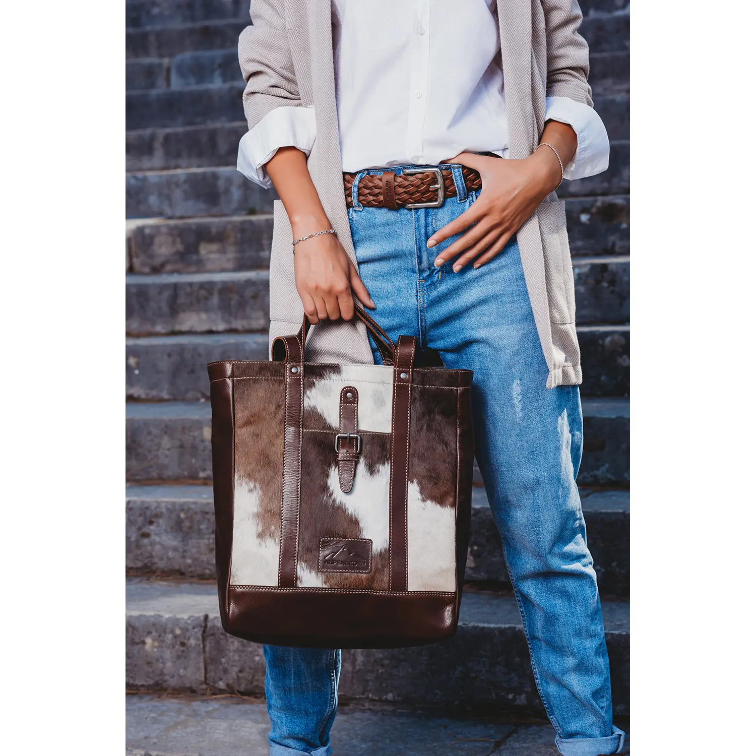 Eine Person in weißem Hemd, beigem Blazer und Blue Jeans hält den Shopper HALLODRI von Alpenleder. Auf einer Steintreppe stehend, liegt der Fokus auf dem lässigen Outfit und dem einzigartigen Kuhfell-Design der Tasche aus braun-weißem Leder.
