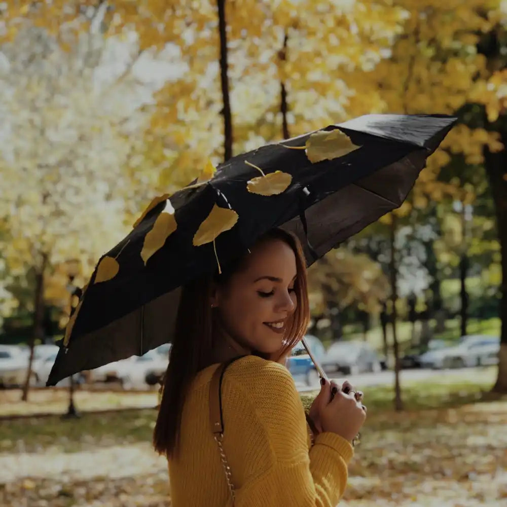 Frau in gelbem Pullover hält einen schwarzen Regenschirm mit gelben Blättern in einem Park.